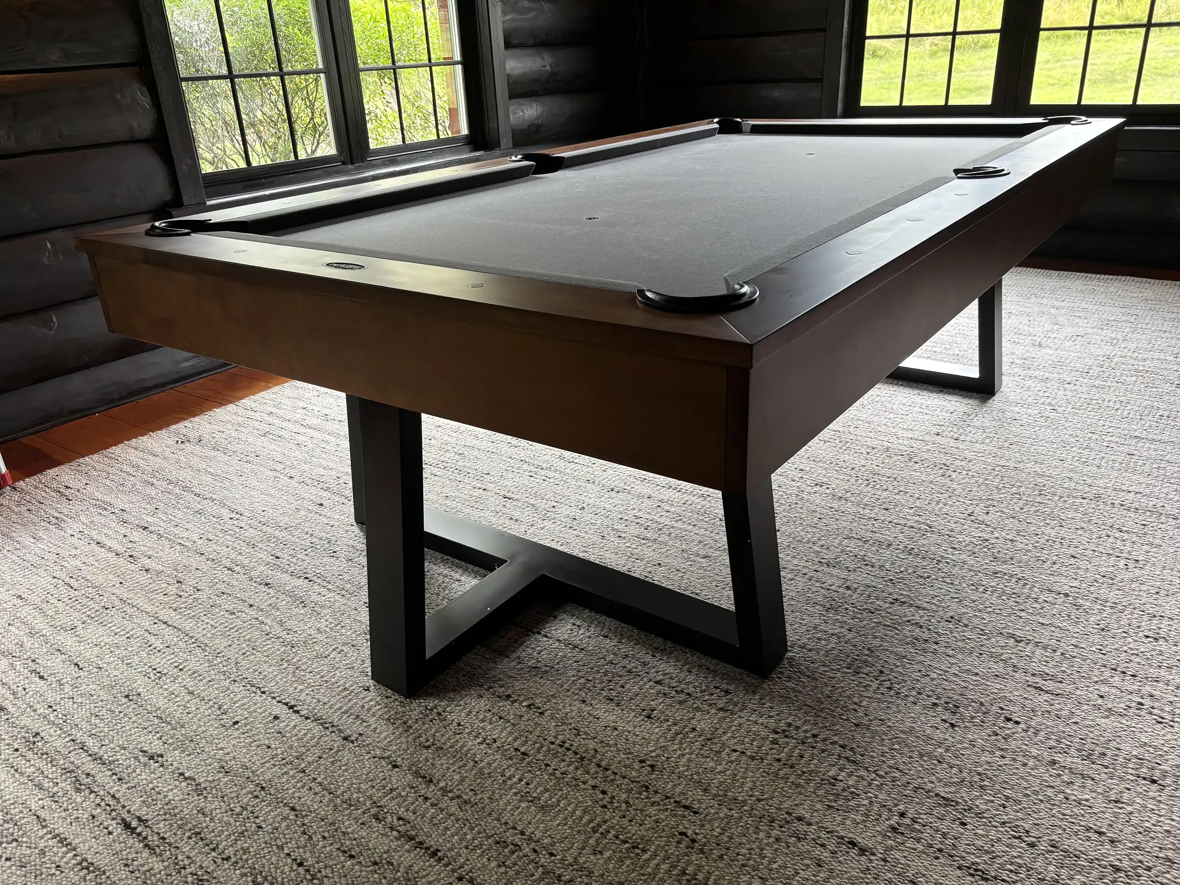 Imperial pool table with walnut finish, grey black felt, and industrial metal legs on a textured rug in a room with large windows.
