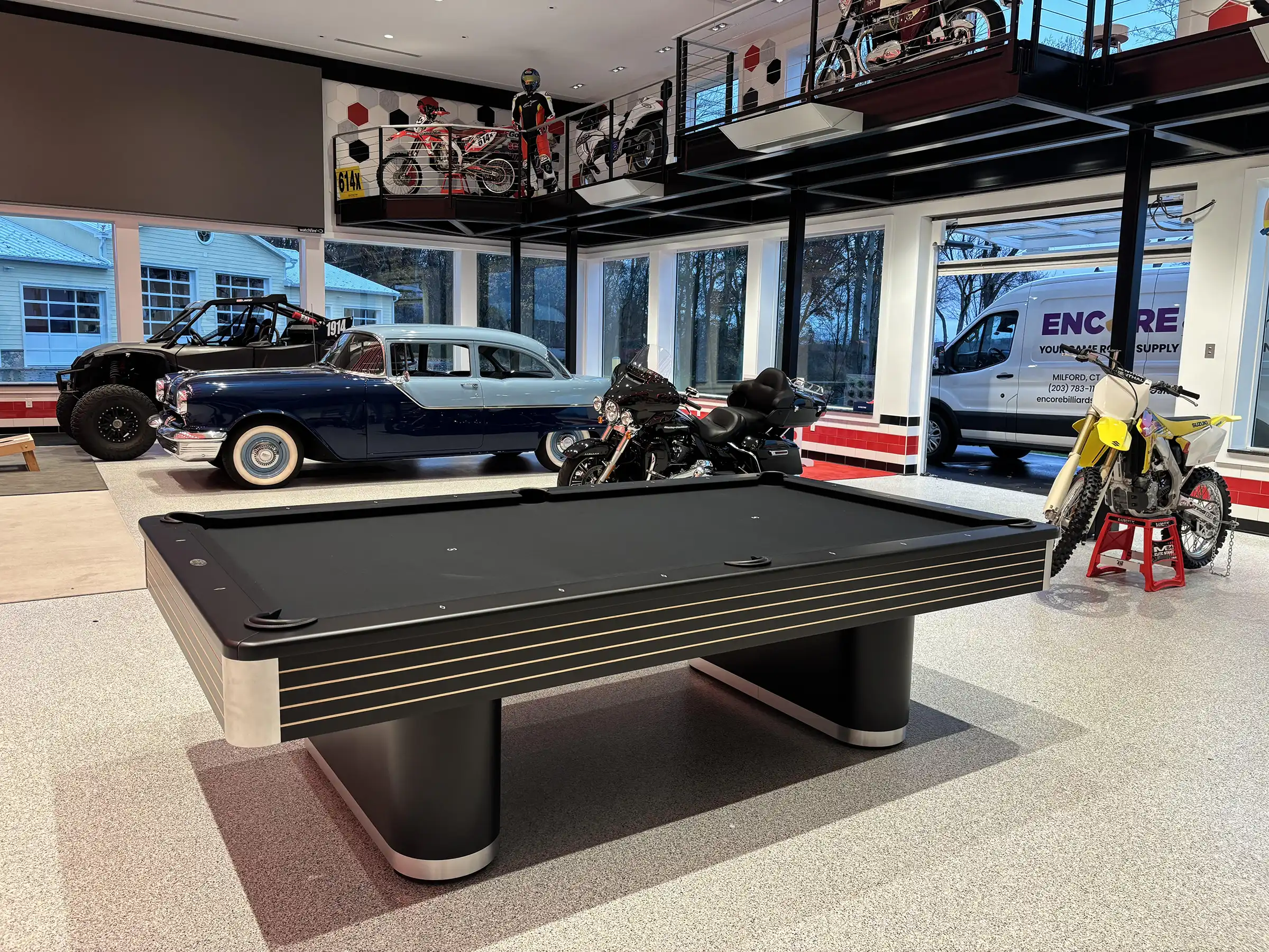 Modern black pool table with gold accents in a luxury garage showroom featuring classic cars, motorcycles, and bright epoxy flooring.