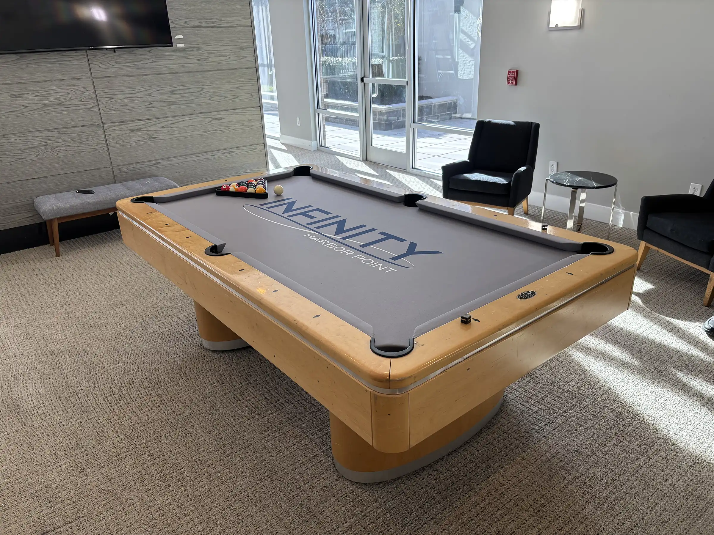 Modern light wood pool table with custom Infinity Harbor Point branding on grey felt, located in a sunny resident amenity lounge area.
