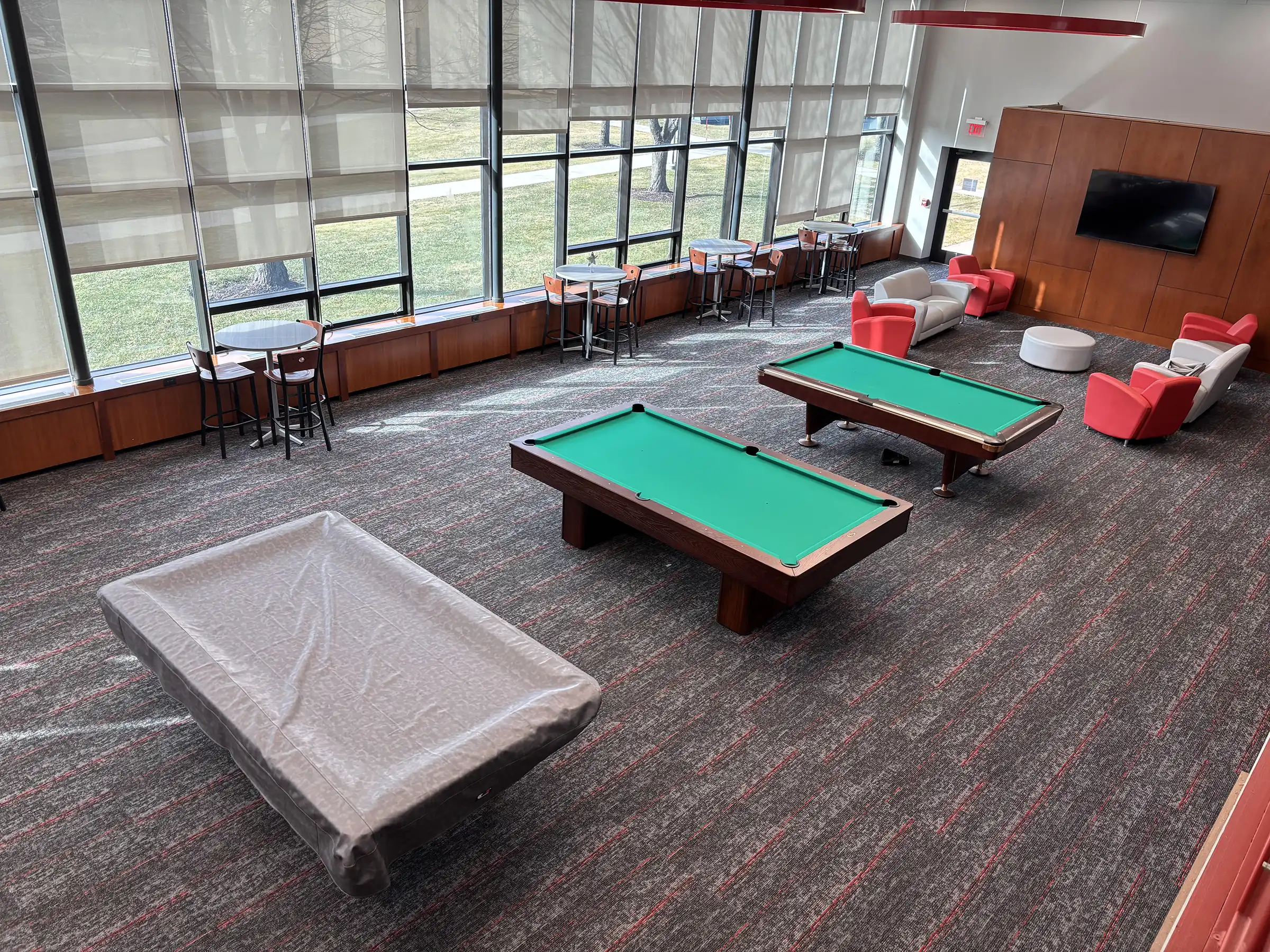 A spacious billiards room at Sacred Heart with three pool tables, red and white lounge seating, high-top cafe tables, and large windows with natural light.