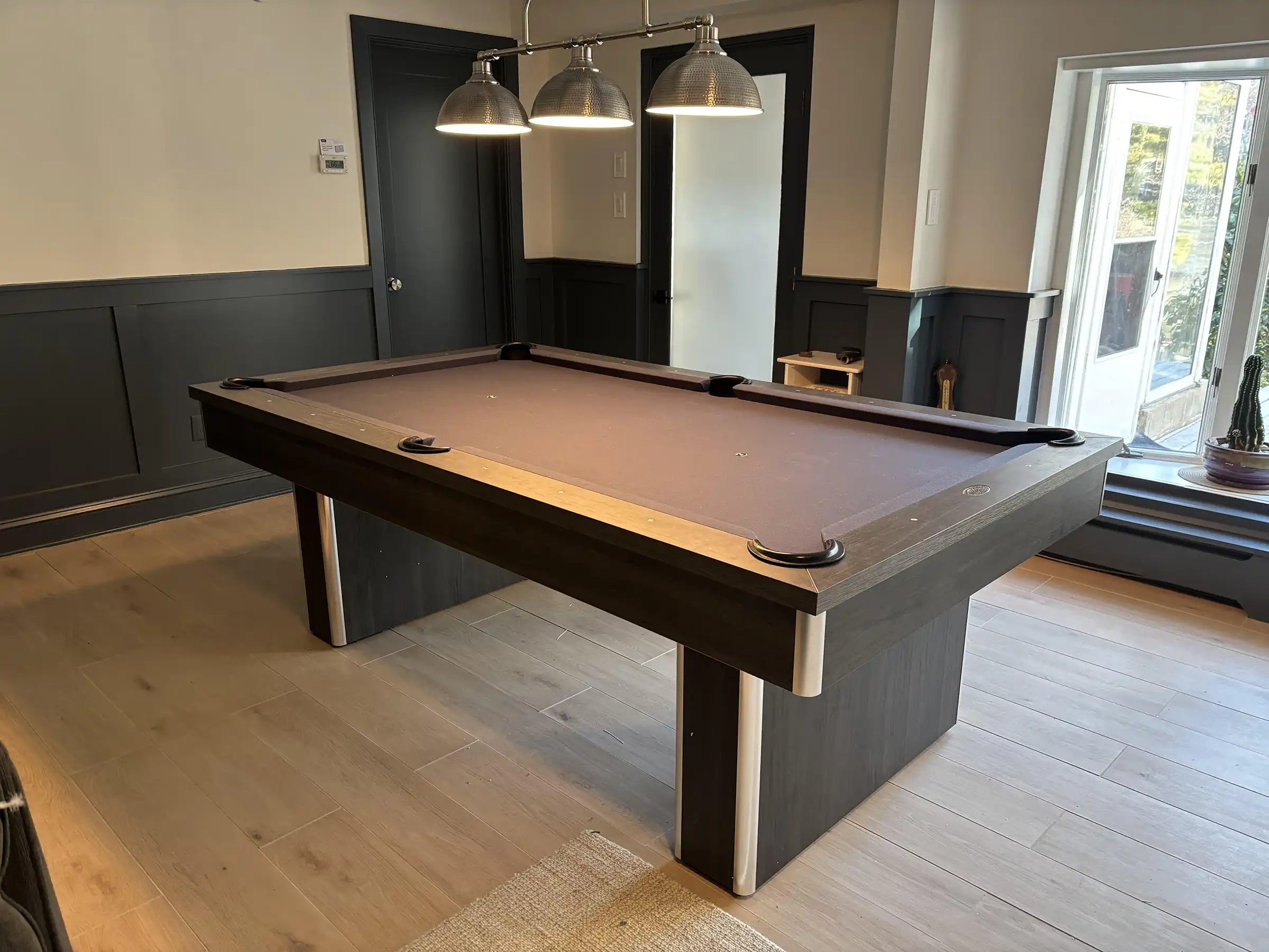 Modern dark wood Olhausen pool table with grey or brown felt in a home game room featuring three industrial pendant lights and light wood flooring.