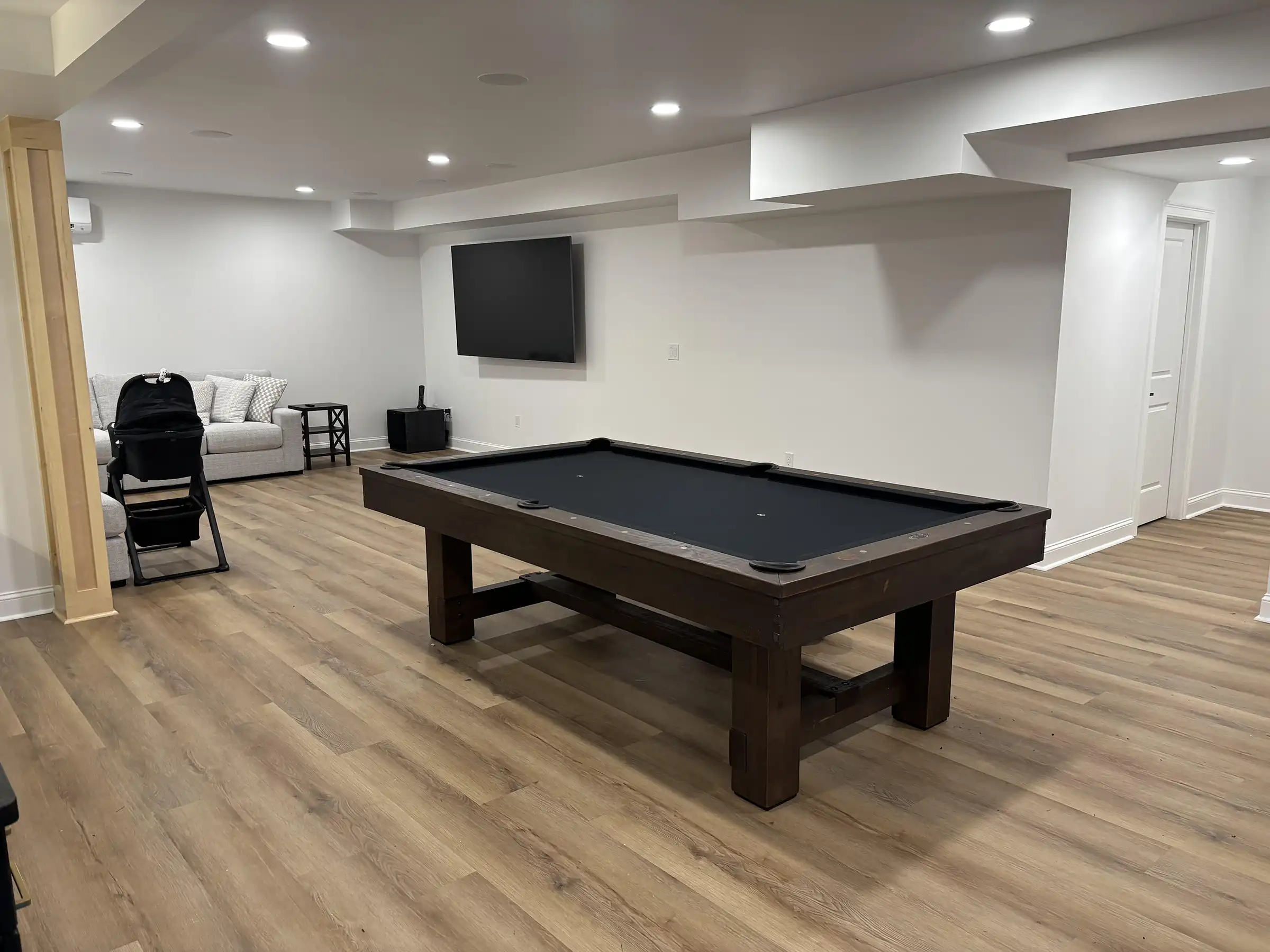 Rustic dark wood Olhausen pool table with sleek black felt in a modern finished basement featuring light wood floors, a wall-mounted TV, and a grey sofa.