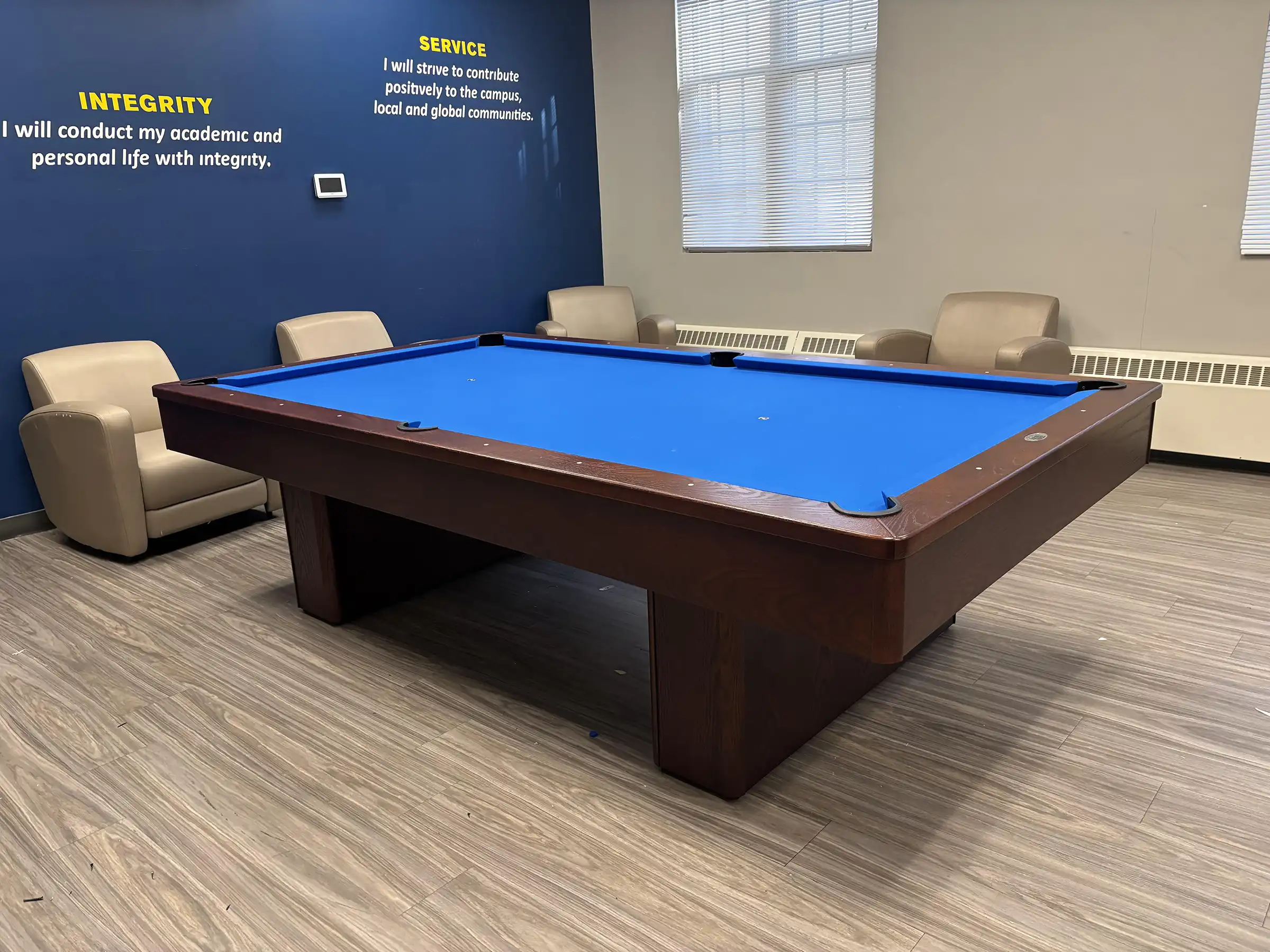 Professional dark wood Olhausen pool table with vibrant blue felt at UNH, featuring tan lounge chairs and a blue accent wall with campus values text.