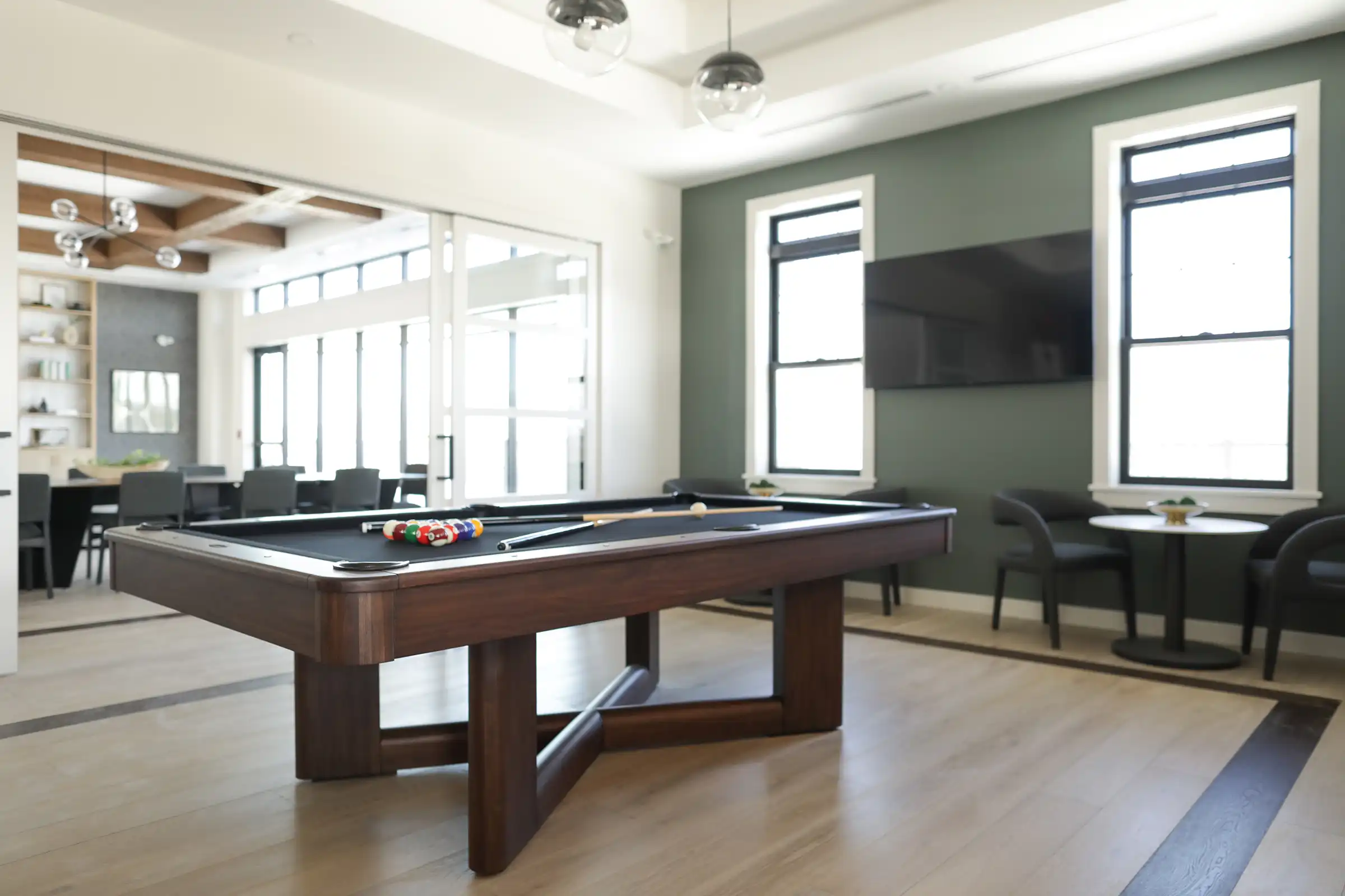 A modern game room featuring a wood pool table with black felt, sage green accent walls, large windows, and an adjacent lounge area with a dining table.
