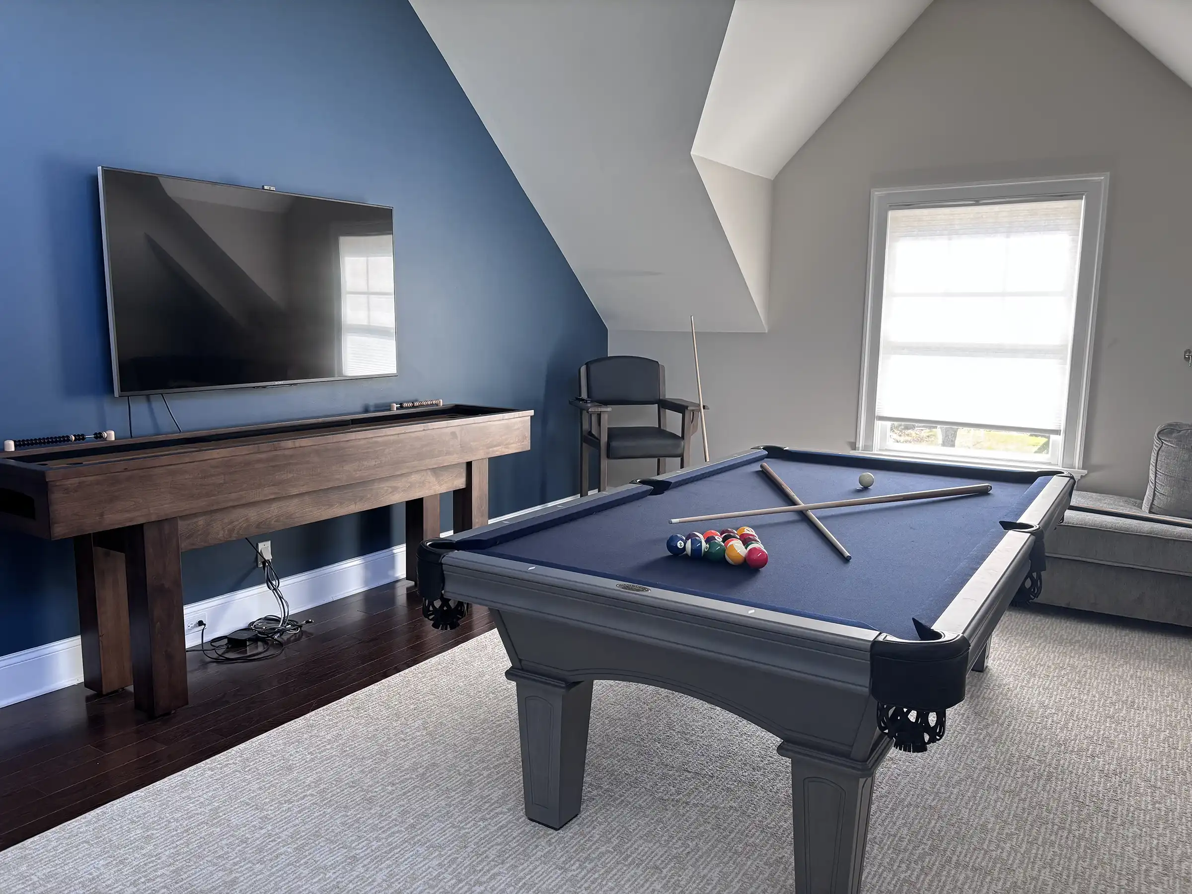 A modern game room featuring a blue felt Olhausen pool table on a light rug, a wooden shuffleboard or bankz or skeech table, and a wall-mounted TV against a navy accent wall.