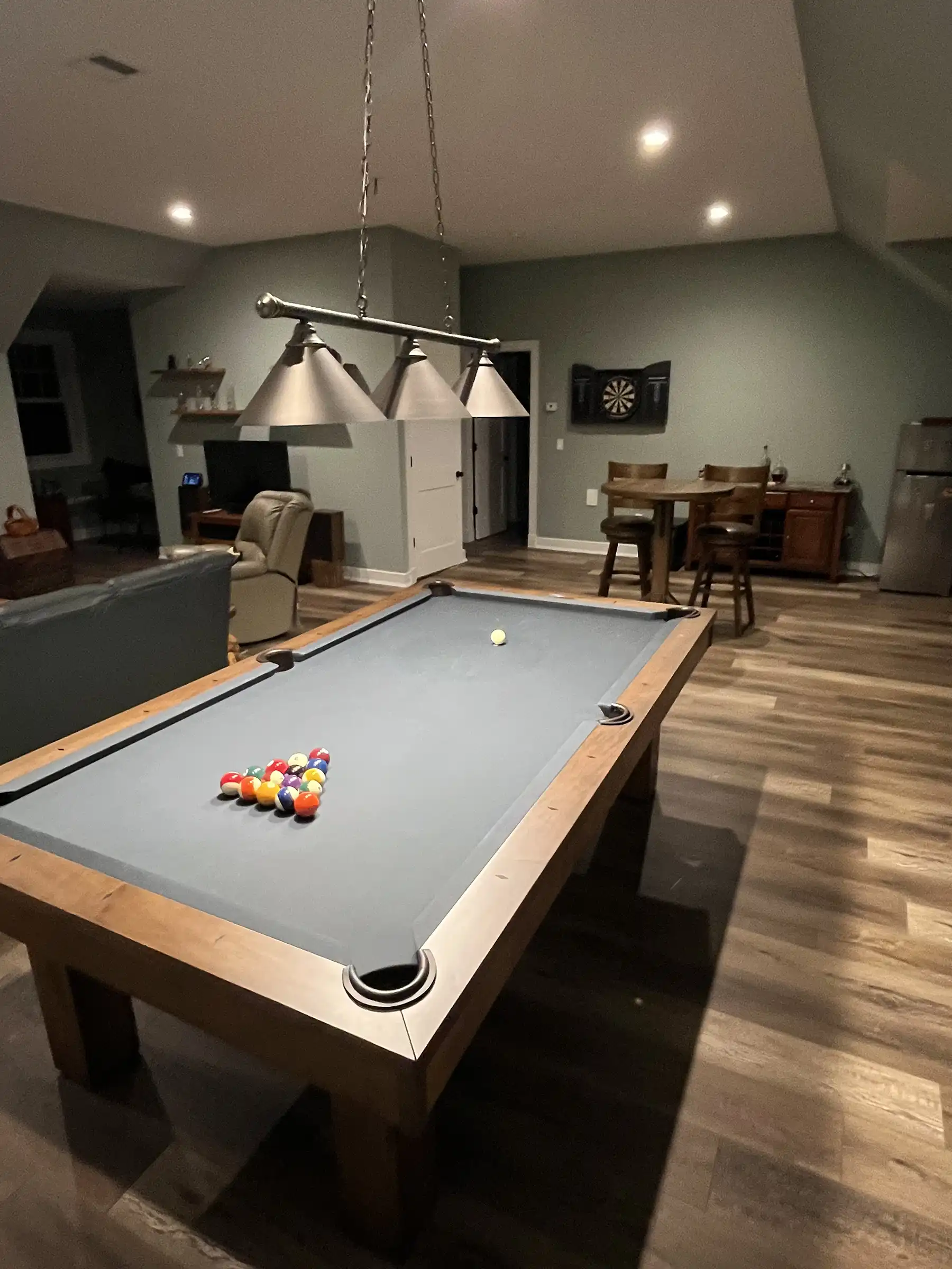 Modern home game room featuring a wood pool table with grey felt, a three-shade hanging light fixture, sage green walls, and a wall-mounted dartboard.