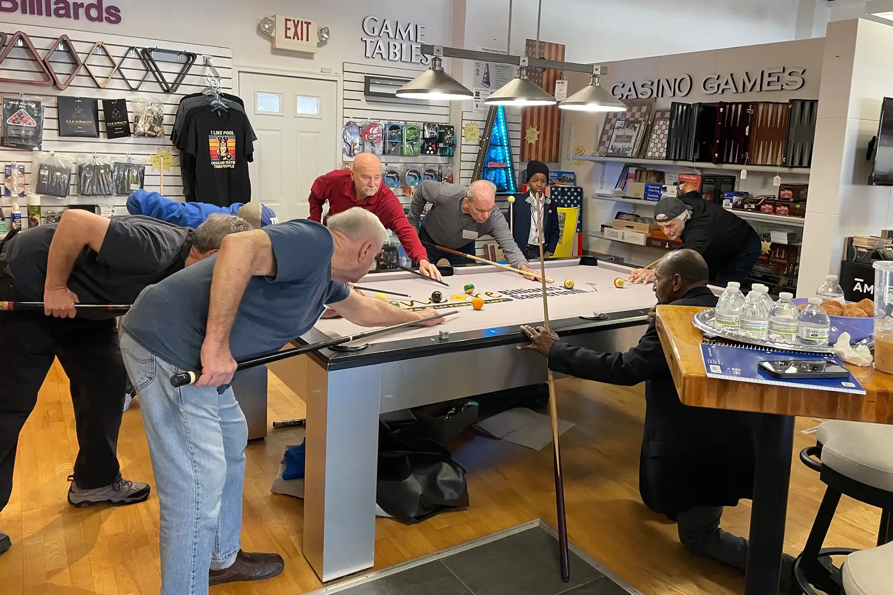 Seven men and a child gather around a sleek, modern pool table in a game room shop, leaning in simultaneously to line up shots with their billiard cues.