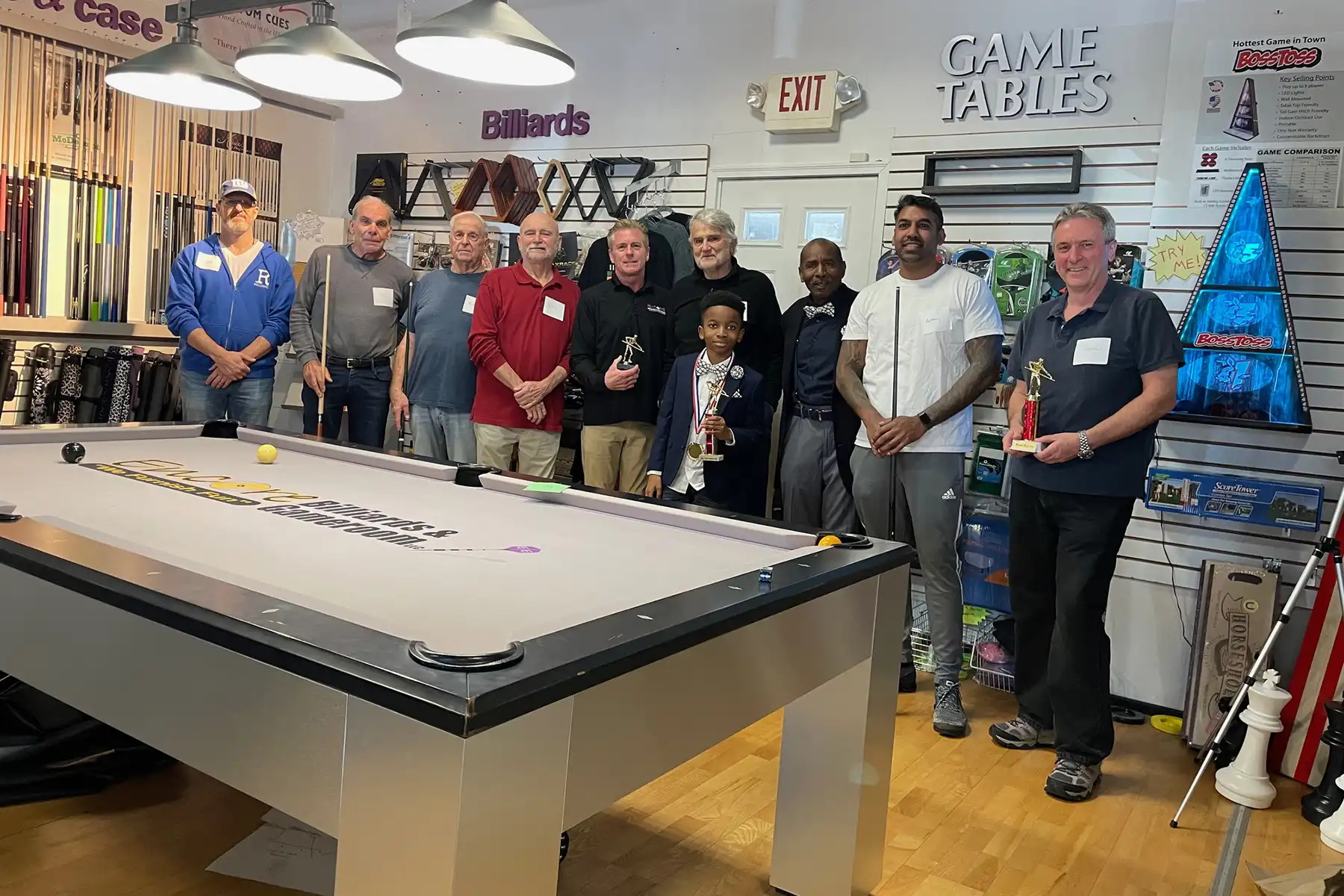 A diverse group of men and a young boy proudly pose with trophies behind a modern billiards table after completing a pool school class in a game store.