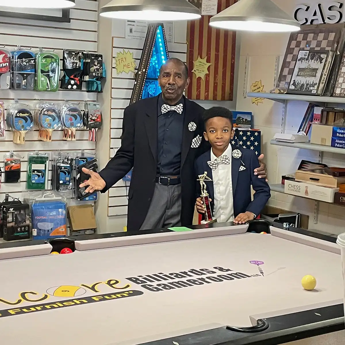 A smartly dressed boy holds a billiards trophy beside a proud man, both wearing bow ties, next to a branded pool table in a game room shop.