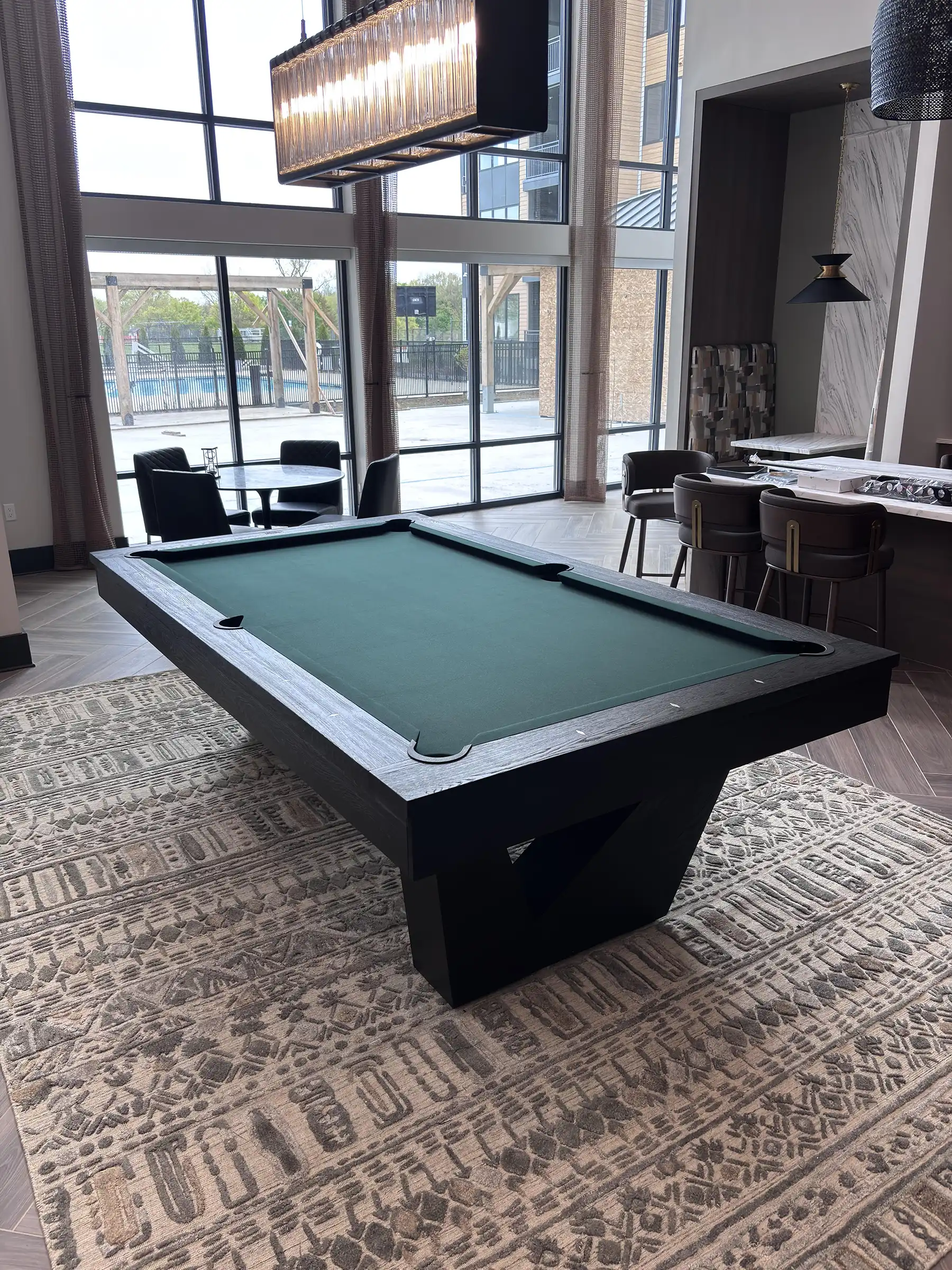 Modern black pool table with green felt on a patterned rug in a bright lounge, featuring large windows, a geometric base, and contemporary lighting.