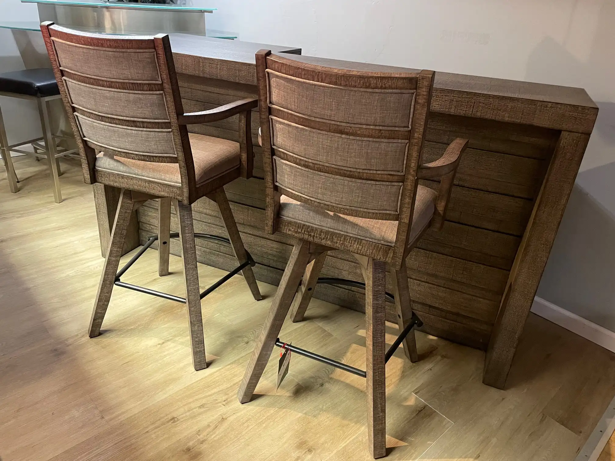 Two upholstered swivel stools from the ECI Furniture Spring Haven 4-piece gameroom bar set, showing the wood slatted backs facing the rustic bar counter.