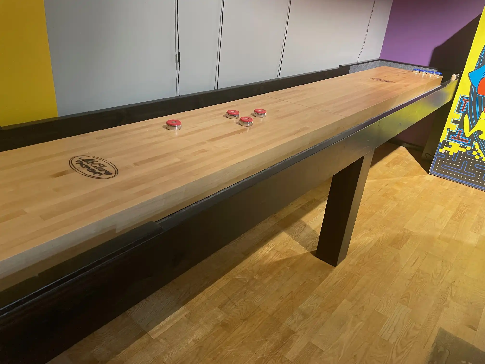 A long McClure shuffleboard table with a light wood surface, black cradle, and red weights on top, located in a game room next to an arcade game.
