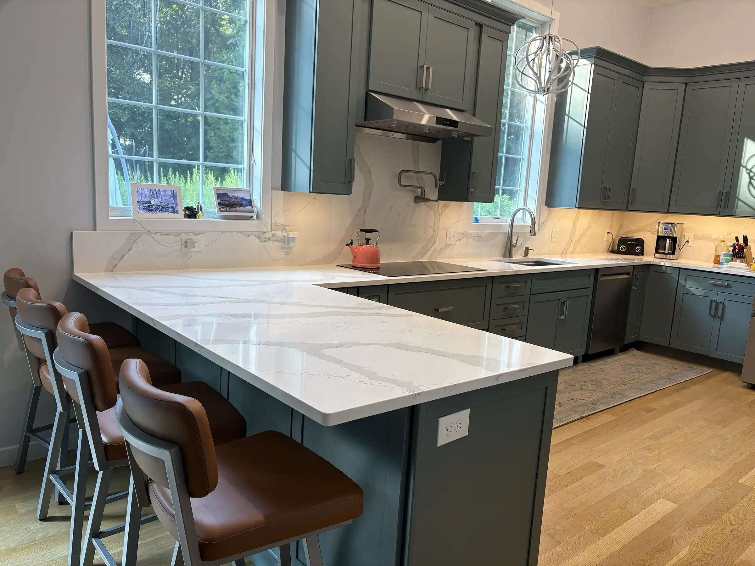 Modern kitchen with dark green cabinets, white veined quartz countertops, stainless steel appliances, and brown leather bar stools at a peninsula counter.