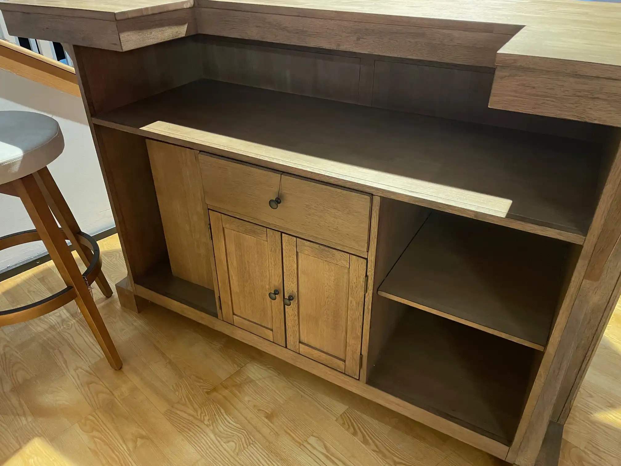 Rear view of a light rustic wood home bar showing storage shelves, cabinet doors, drawers, and a backless bartender stool.