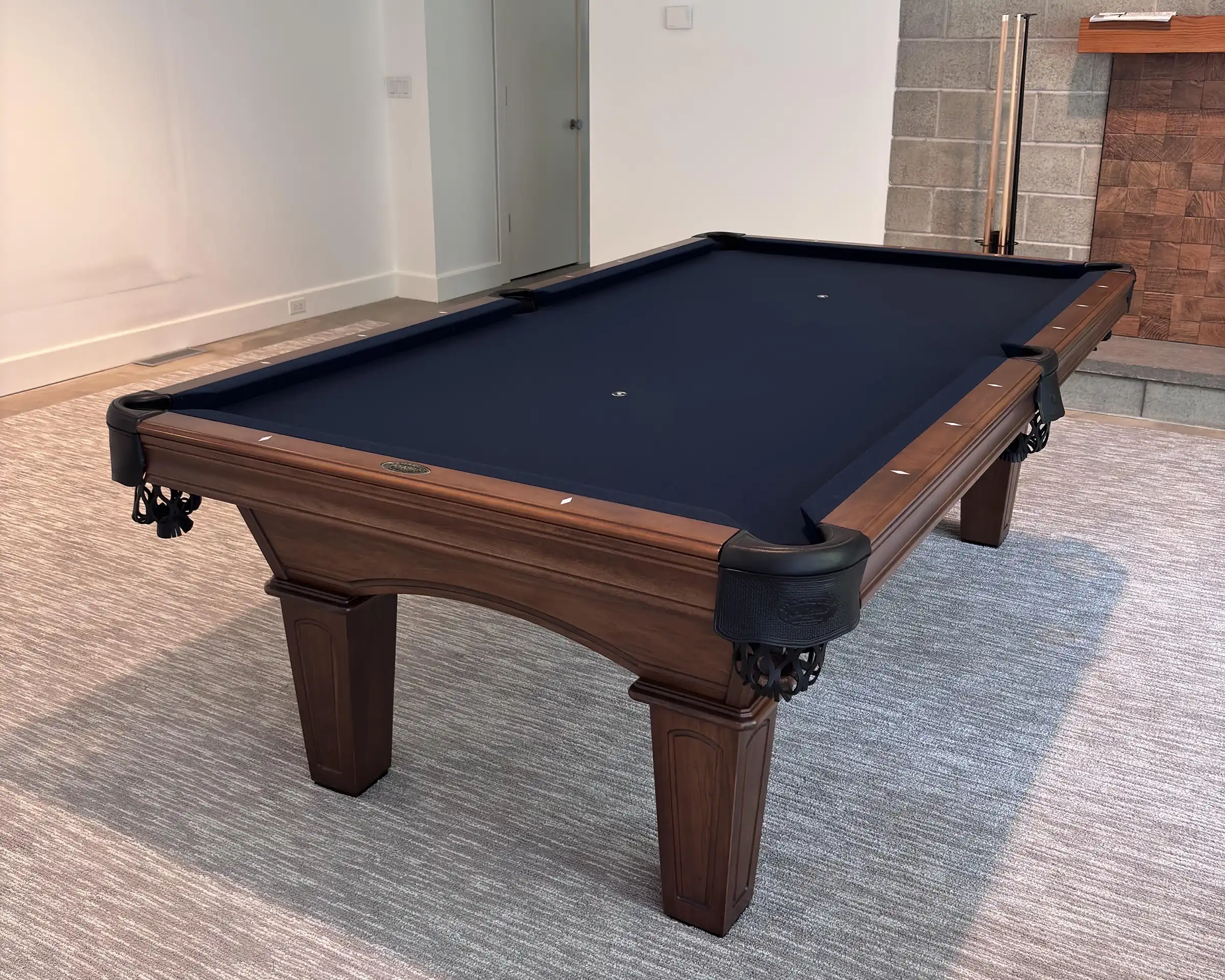 Classic Olhausen wood pool table featuring navy blue felt, leather drop pockets, and tapered legs set on a neutral area rug in a modern room.