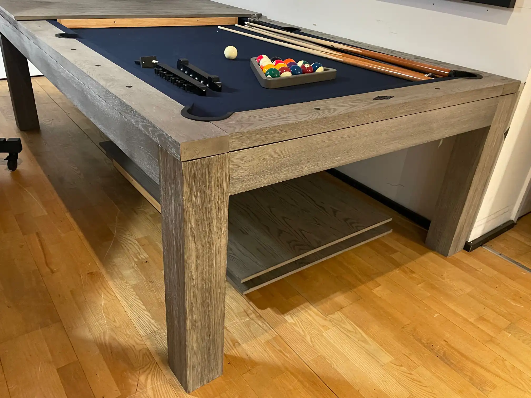 Brunswick Sanibel rustic grey pool table featuring blue felt cloth, pool cues, a racked set of billiard balls, and dining top storage underneath.