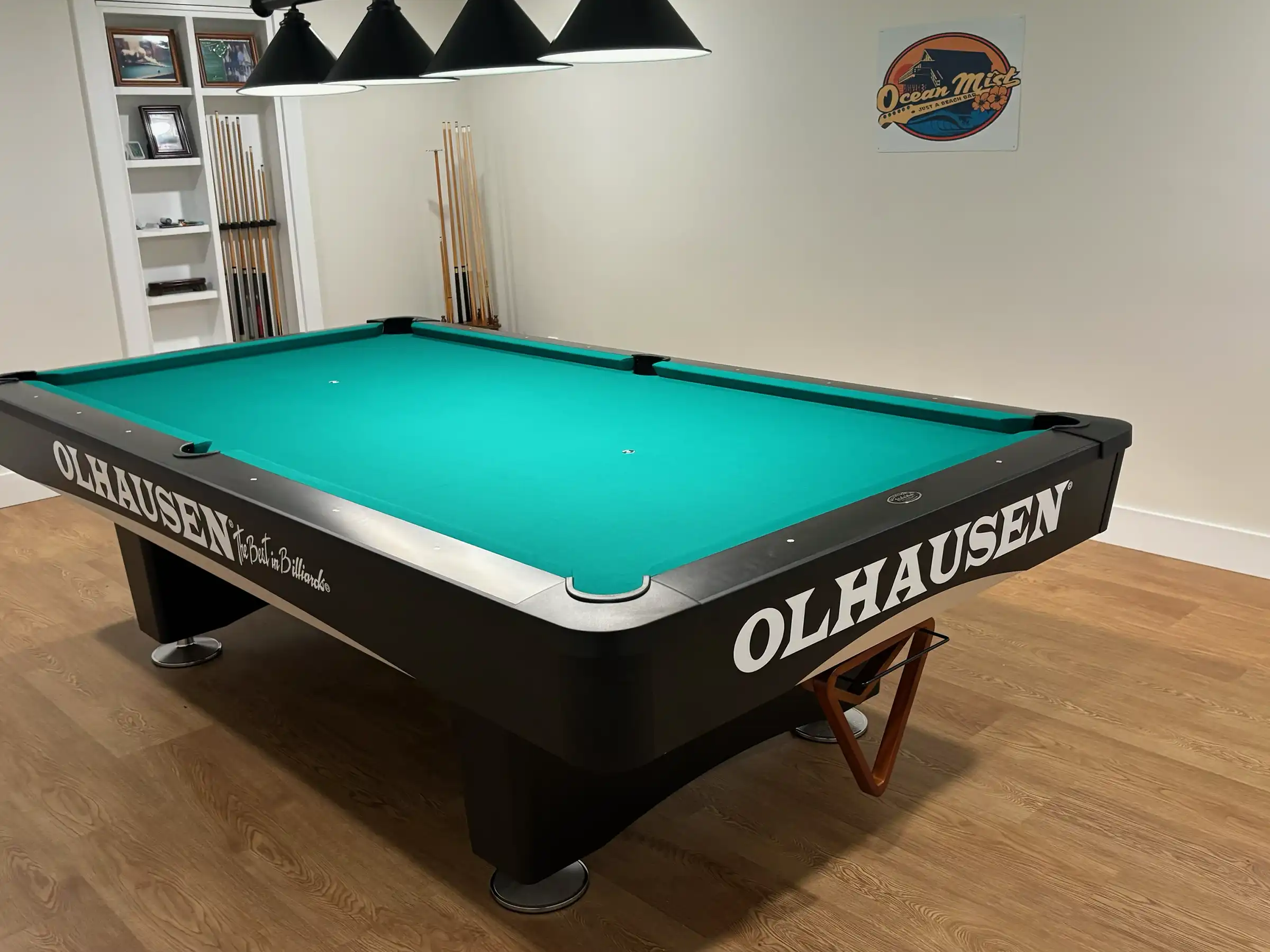 Black Olhausen pool table with vibrant green felt in a modern game room, featuring hardwood floors, overhead hanging lights, and wooden cue sticks.