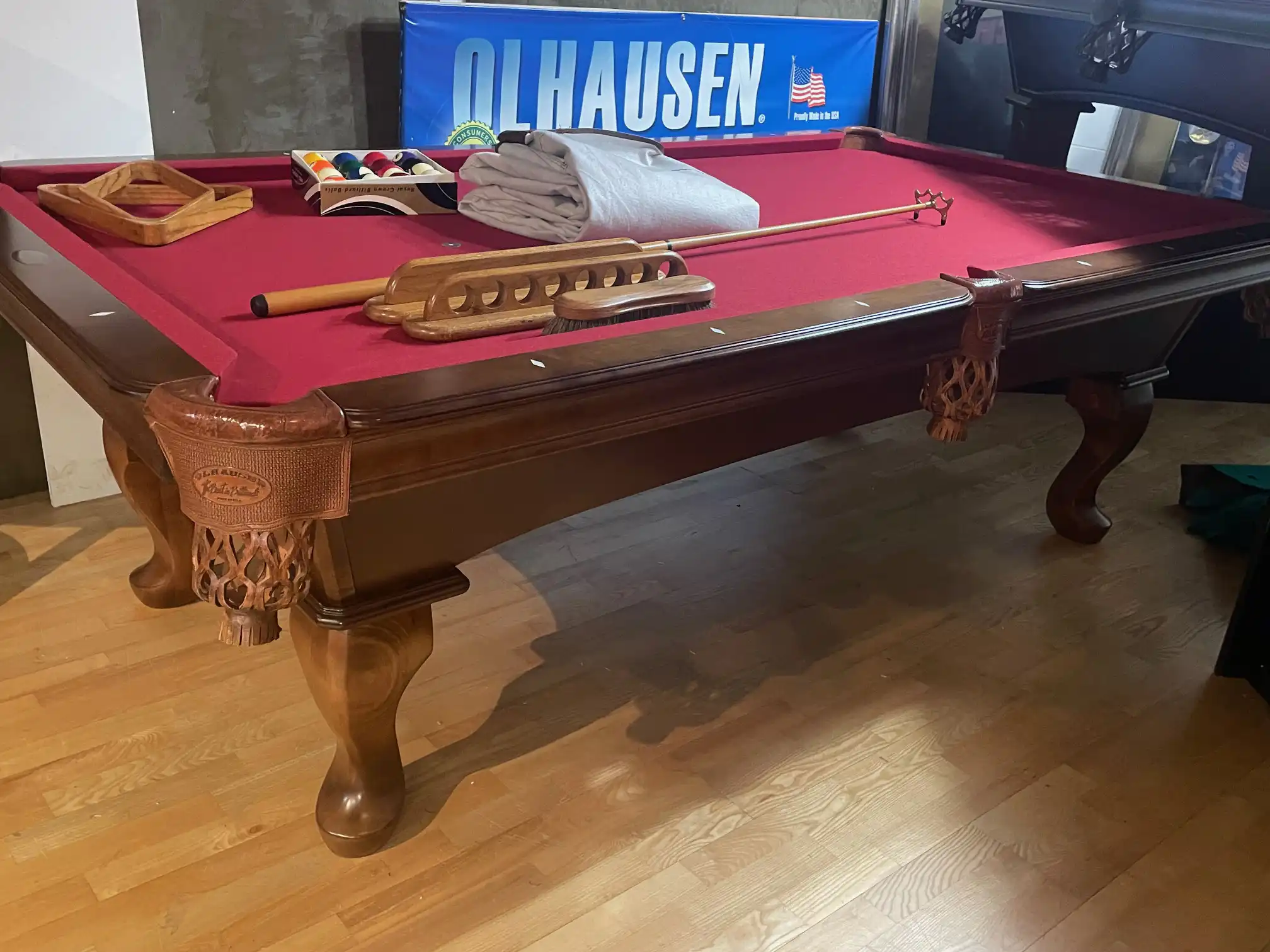 Olhausen wooden pool table with red felt, leather drop pockets, and billiard accessories including balls, cues, and a rack.