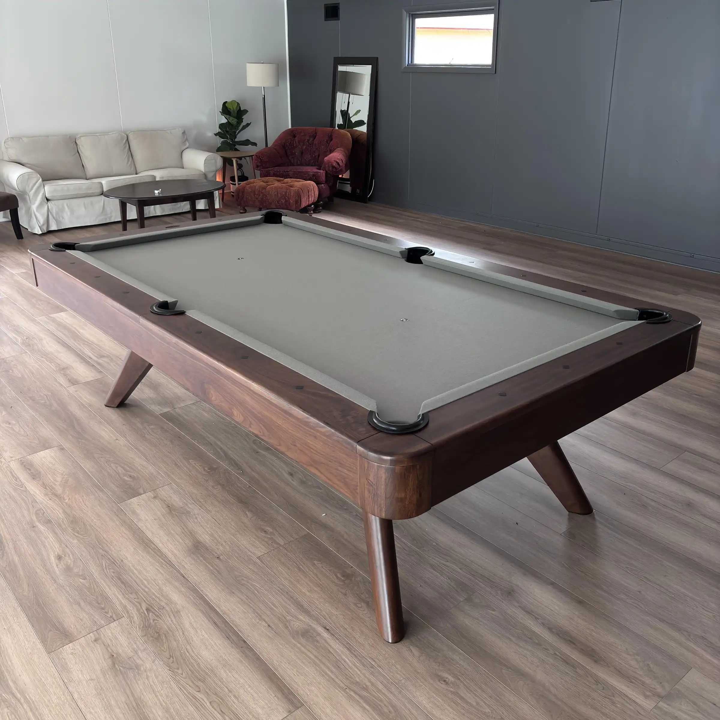 Mid-century modern walnut pool table with grey felt on hardwood flooring in a contemporary game room setting beside lounge furniture.