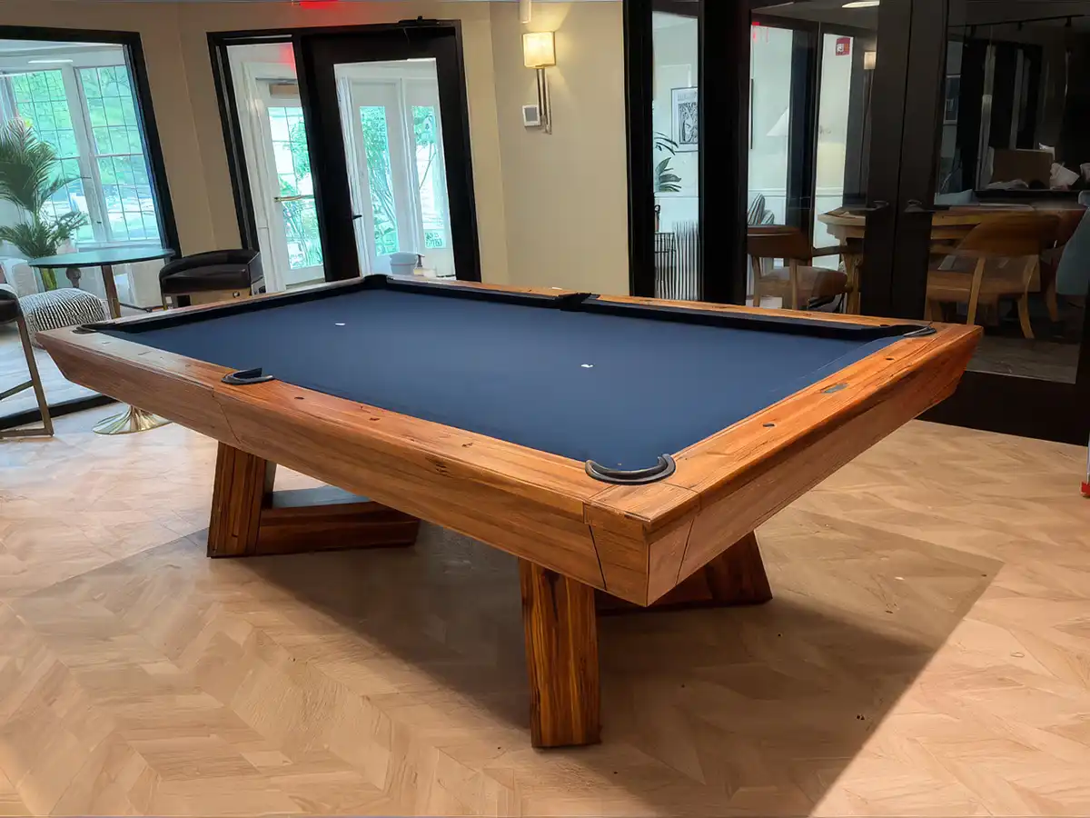 Rustic modern wooden pool table with navy blue felt cloth set on a herringbone hardwood floor inside a bright contemporary game room lounge.