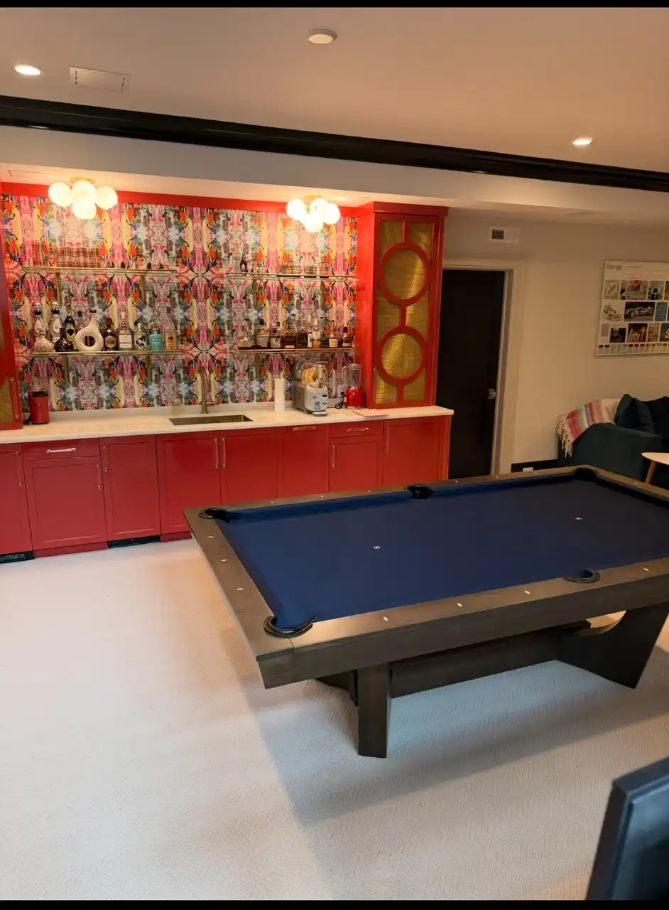 Modern dark wood pool table with navy blue felt set in a stylish basement game room featuring a bold red wet bar and colorful patterned wallpaper.