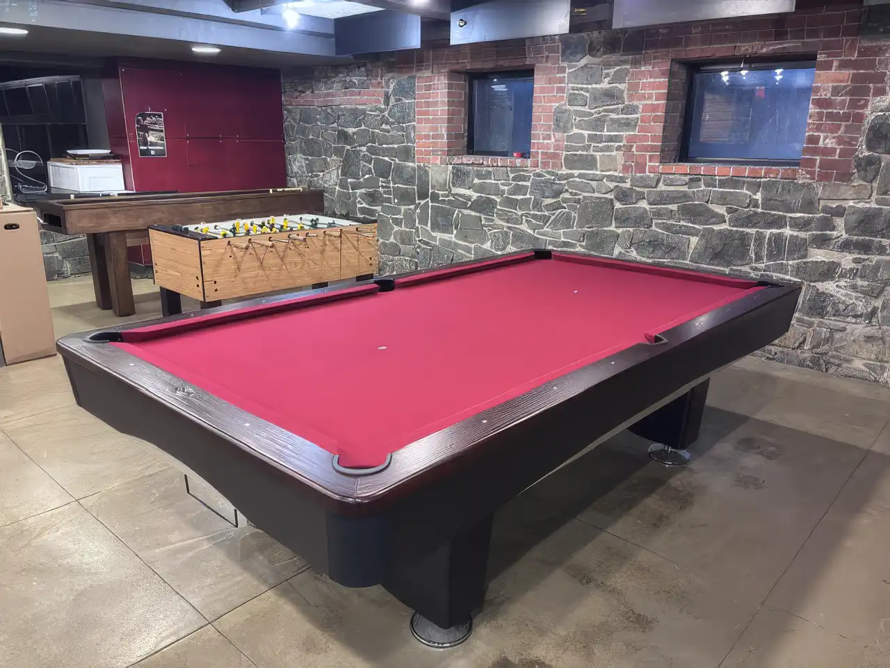 Alt Text: Modern billiard table with red felt and dark wood finish in a basement game room featuring an exposed stone wall, foosball table.