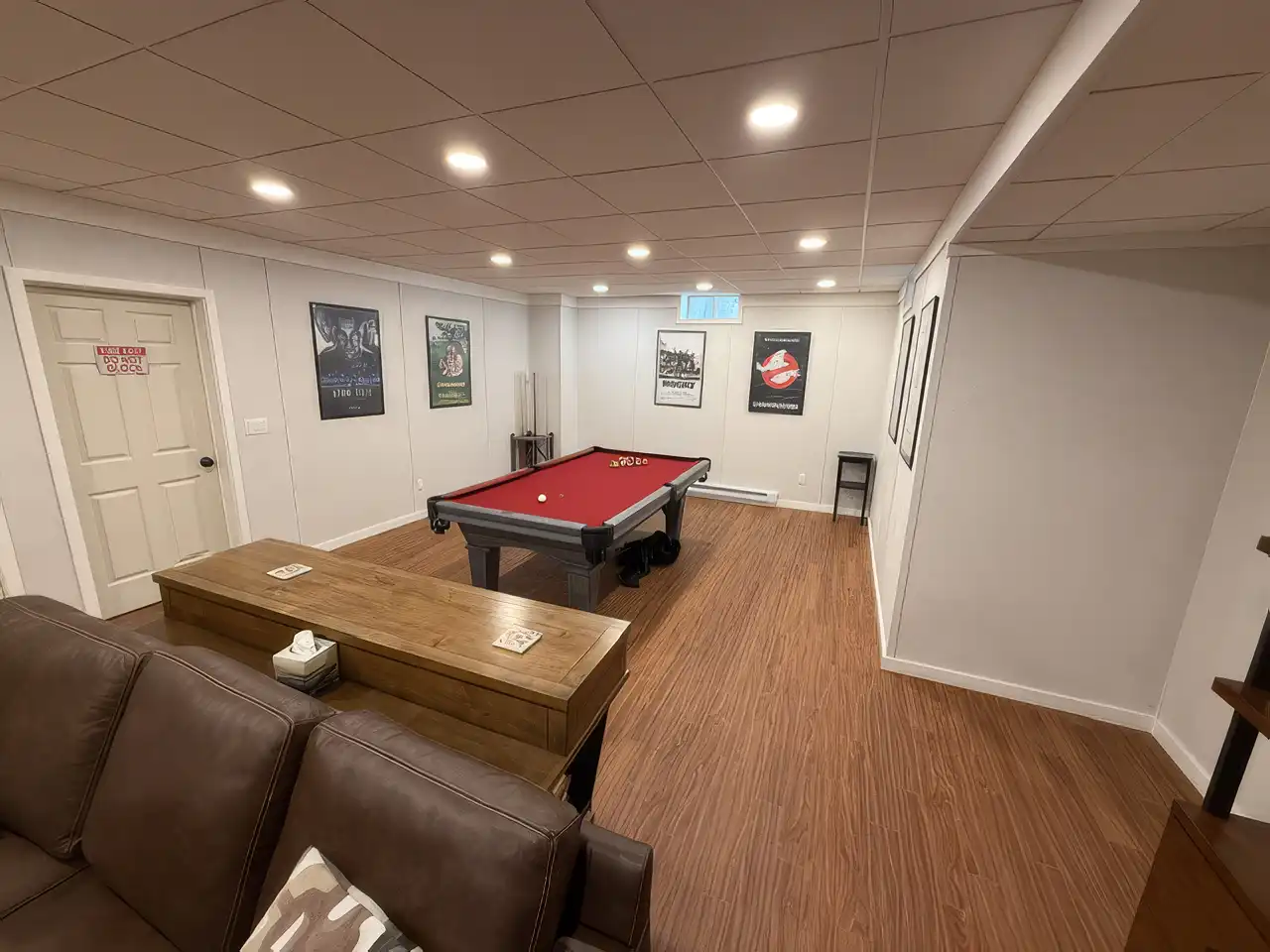 Finished basement game room featuring a red felt pool table, wood laminate flooring, framed movie posters on the wall, and modern recessed ceiling lights.