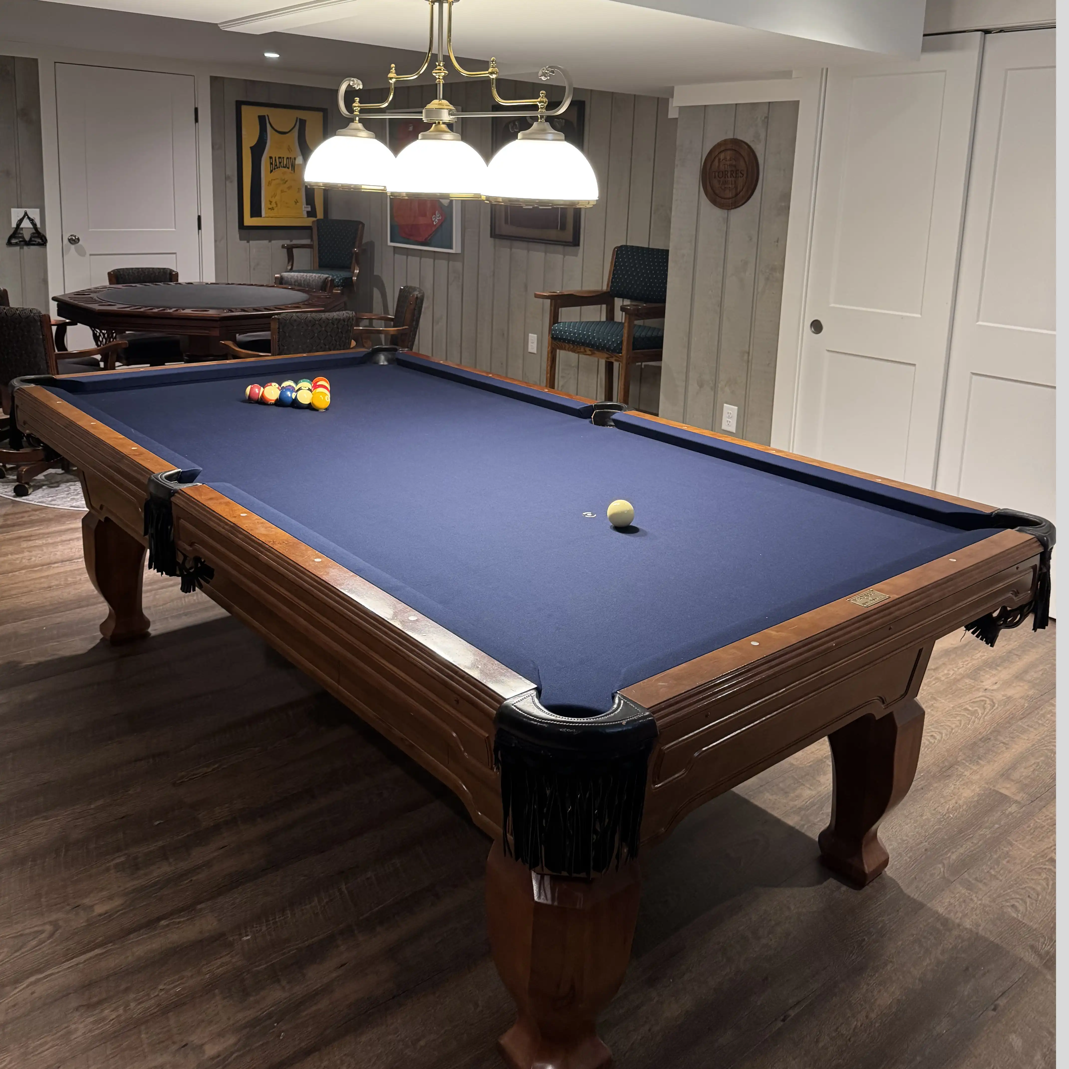 Classic wooden pool table with blue felt and fringe pockets illuminated by a brass chandelier in a finished basement game room.