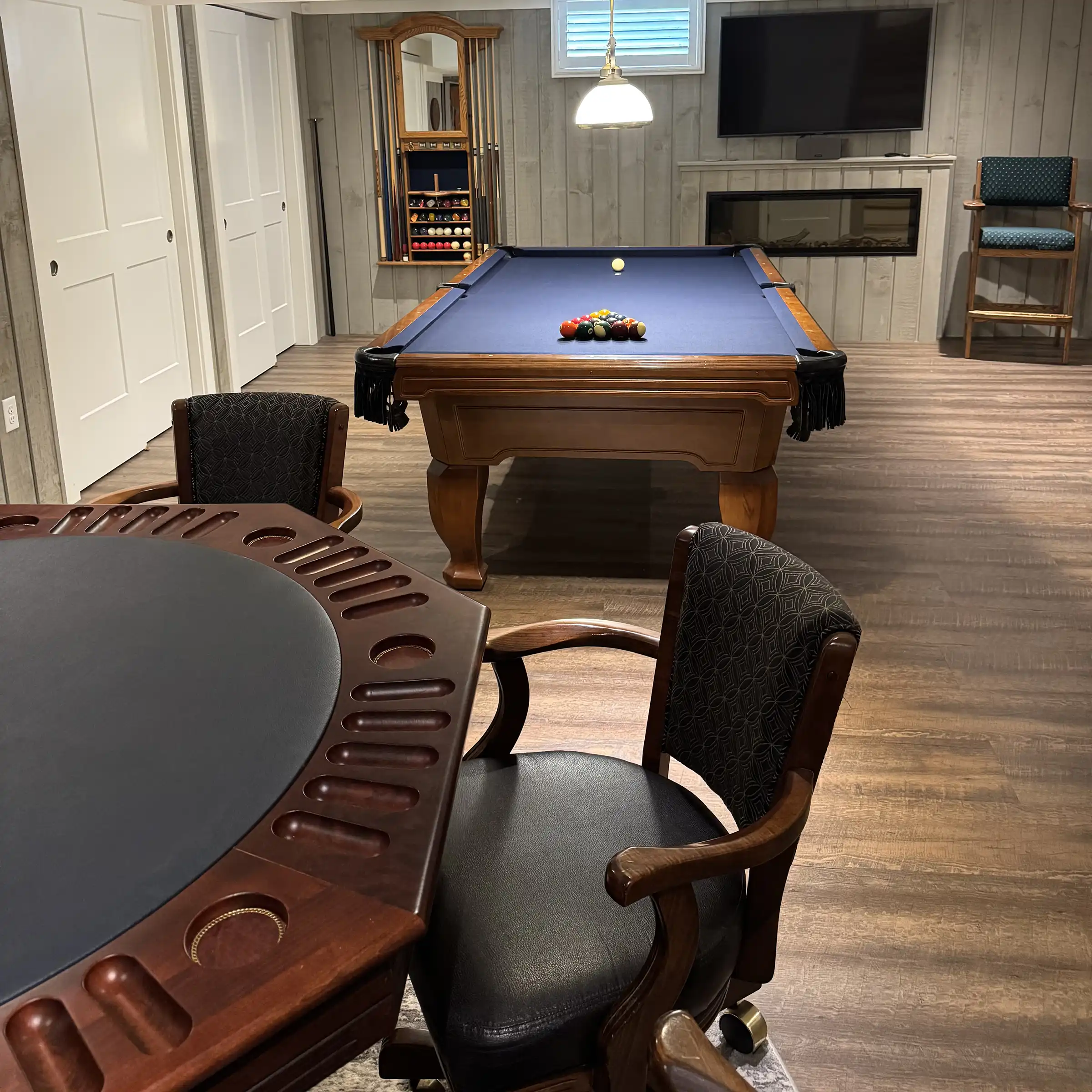 Finished game room featuring an octagonal wooden poker table with leather armchairs in the foreground and a pool table with blue felt in the back.
