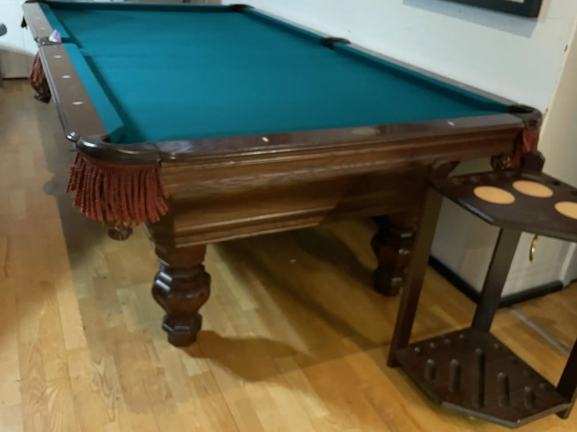 Olhausen Wentworth pool table featuring green felt, fringed drop pockets, and carved solid wood legs, placed beside a matching cue rack in a game room.