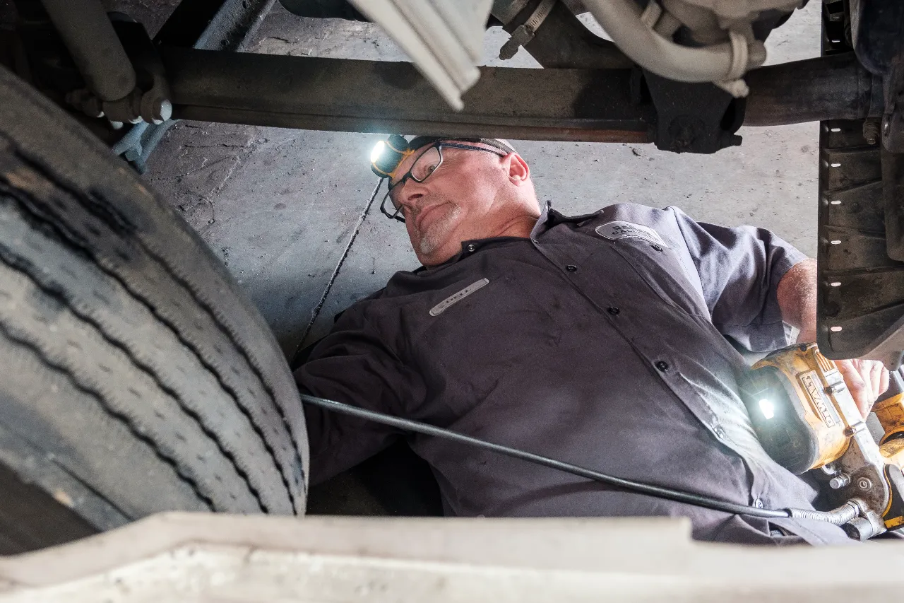 Mechanic lying under truck chassis with headlamp, inspecting components near front wheel.