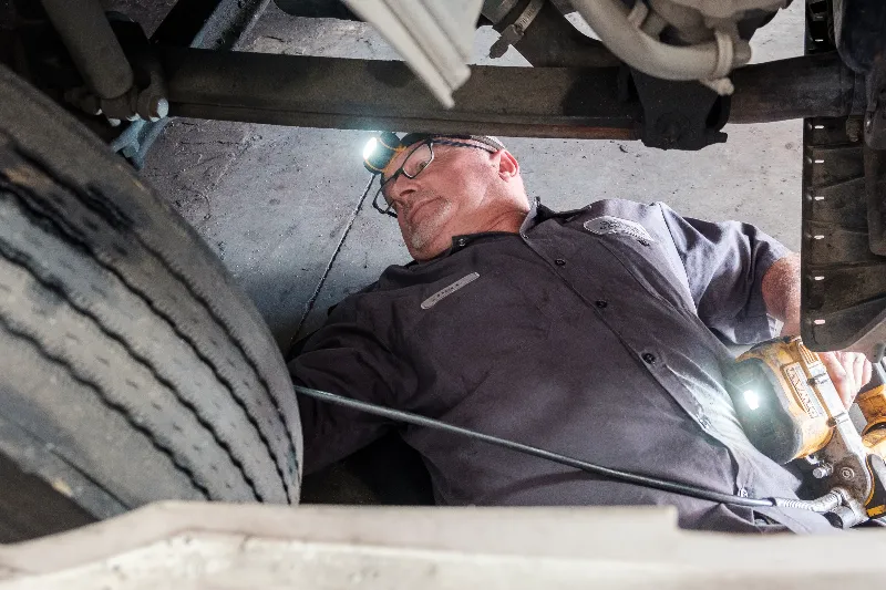 Mechanic lying under truck chassis with headlamp, inspecting components near front wheel.