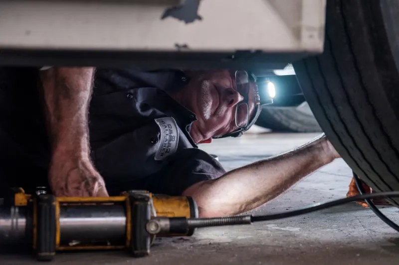Heavy-duty truck repair technician works beneath a vehicle using tools, wearing a headlamp and a B & H Gallagher uniform patch.