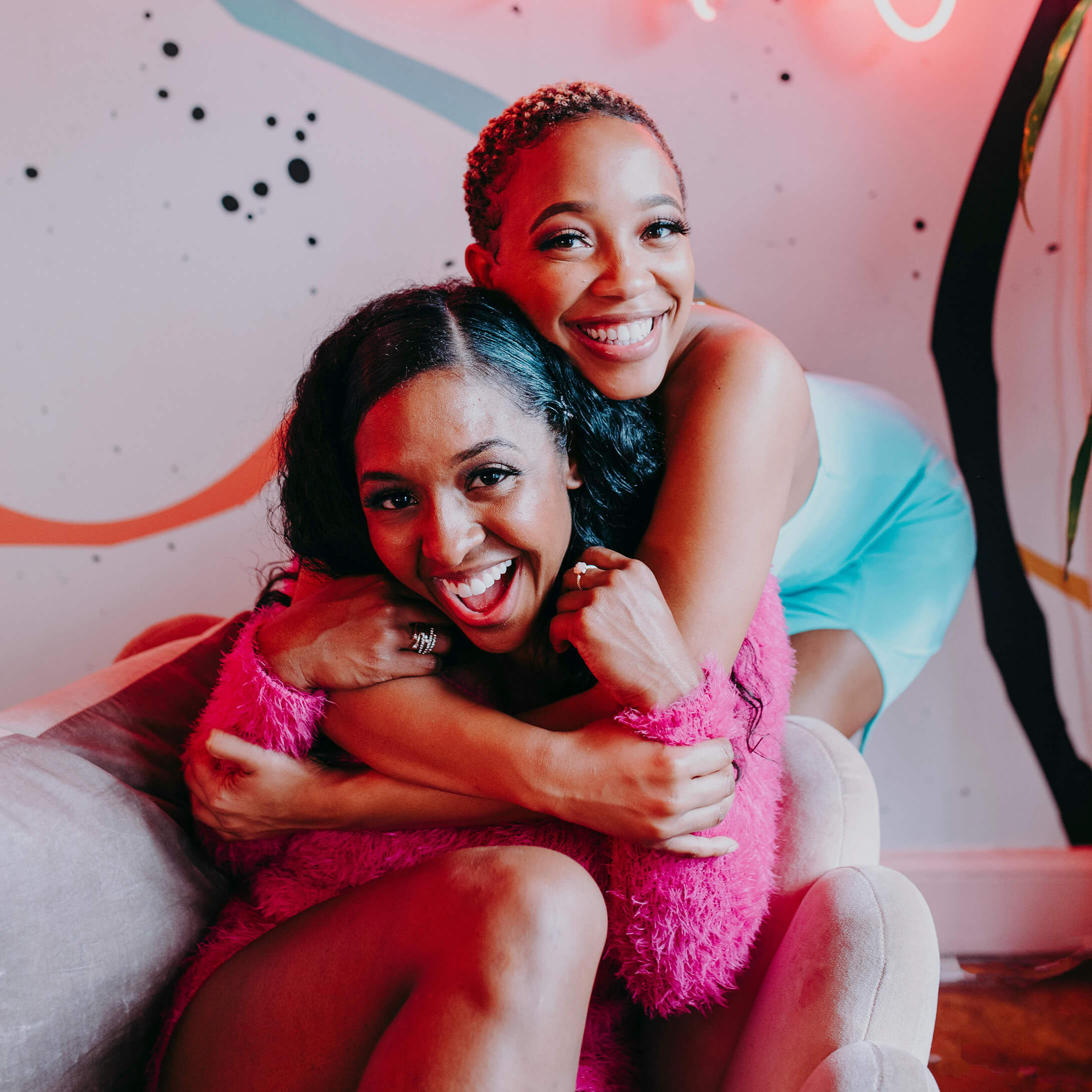 Two women smiling and hugging on a couch with colorful background and soft pink fuzzy blanket.