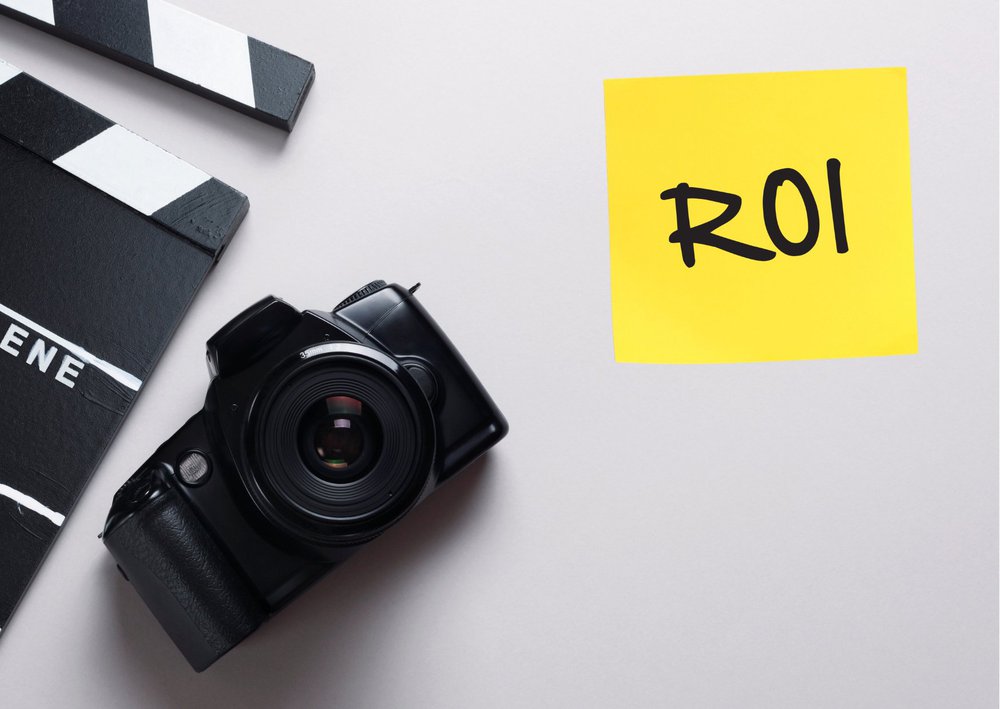 A DSLR camera and movie clapperboard placed beside a yellow sticky note with "ROI" written on it against a white background.