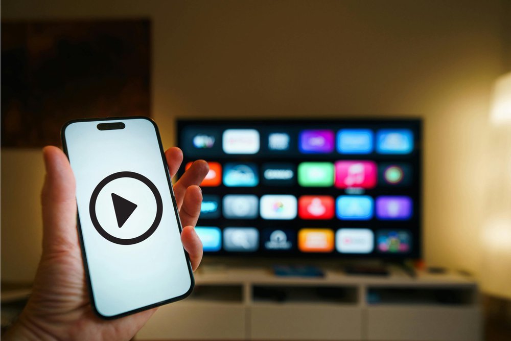 A hand holding a smartphone displaying a play button icon with a smart TV showing streaming app icons in the background.
