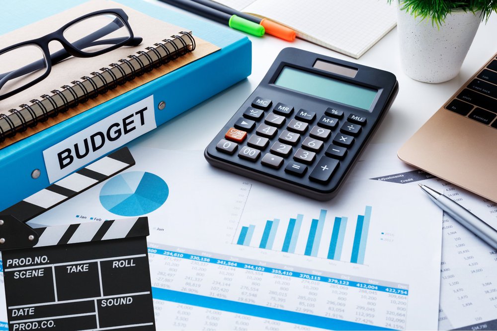 A desk with a movie clapperboard, calculator, budget binder, financial charts, and spreadsheets representing video production budget planning.