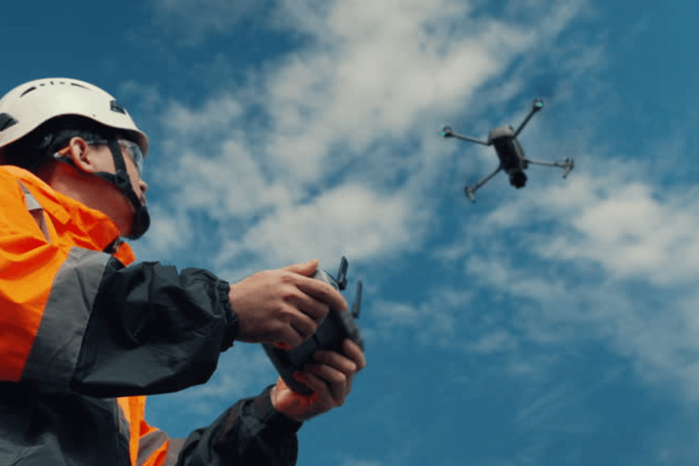 A worker in a hard hat and safety gear pilots a drone with a remote control against a blue sky.