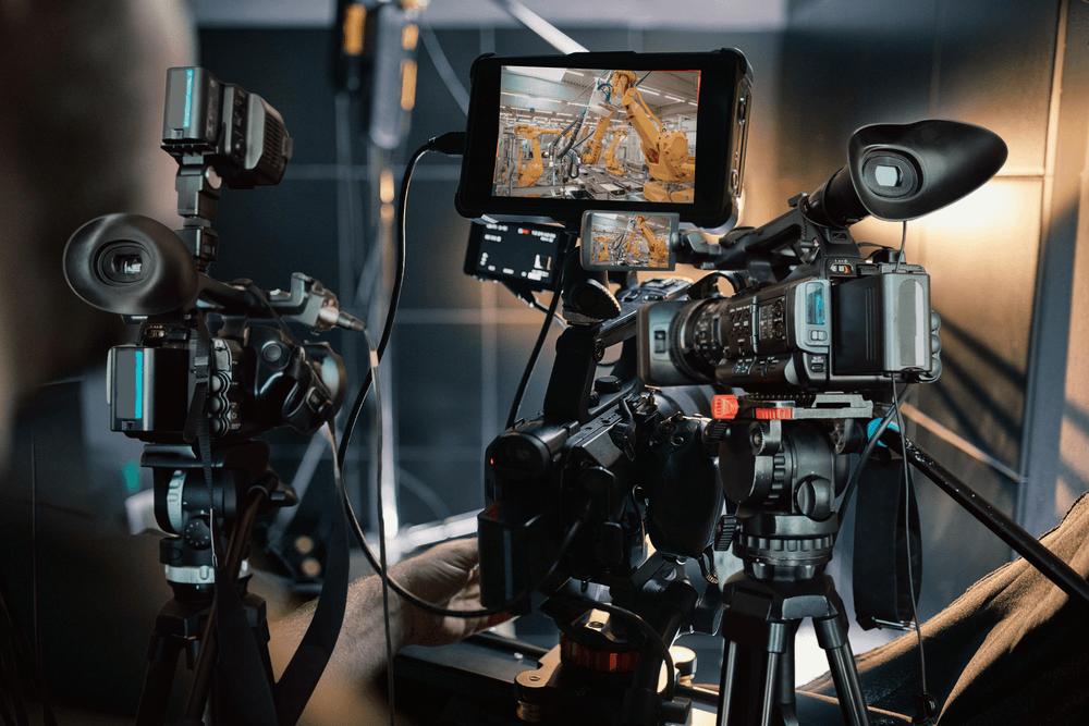 Video cameras on tripods with a monitor showing robotic arms on an automated production line.