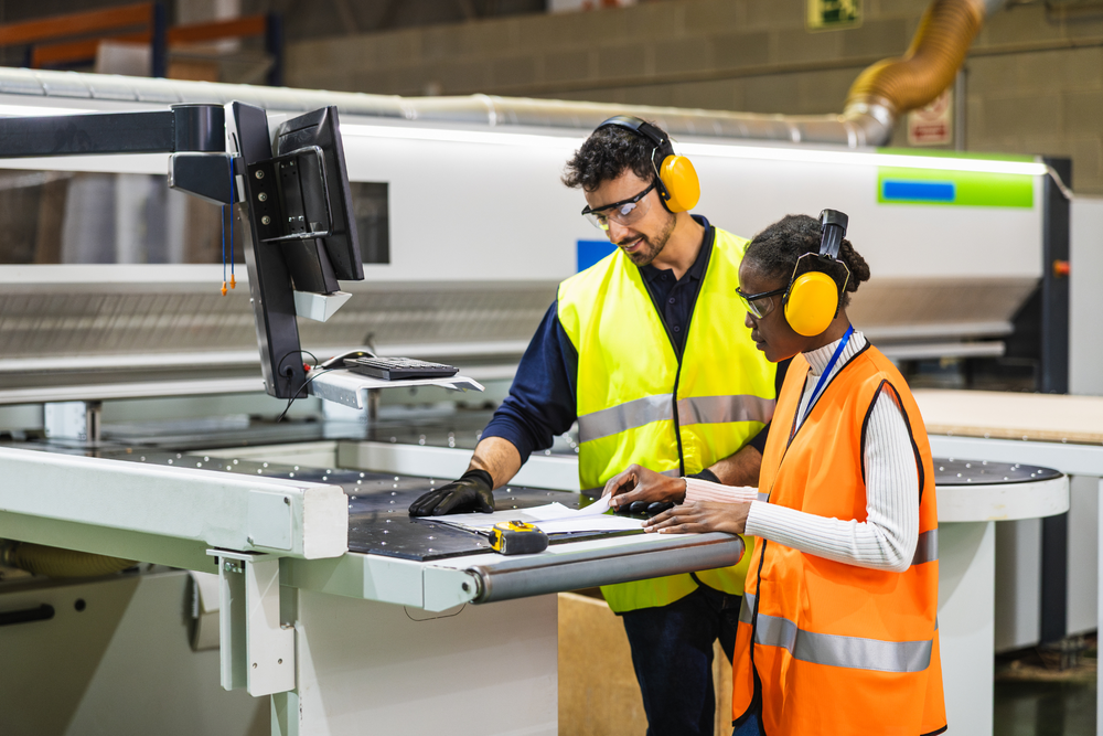 Two workers in high-visibility vests review a document beside CNC panel-cutting equipment in a manufacturing facility.