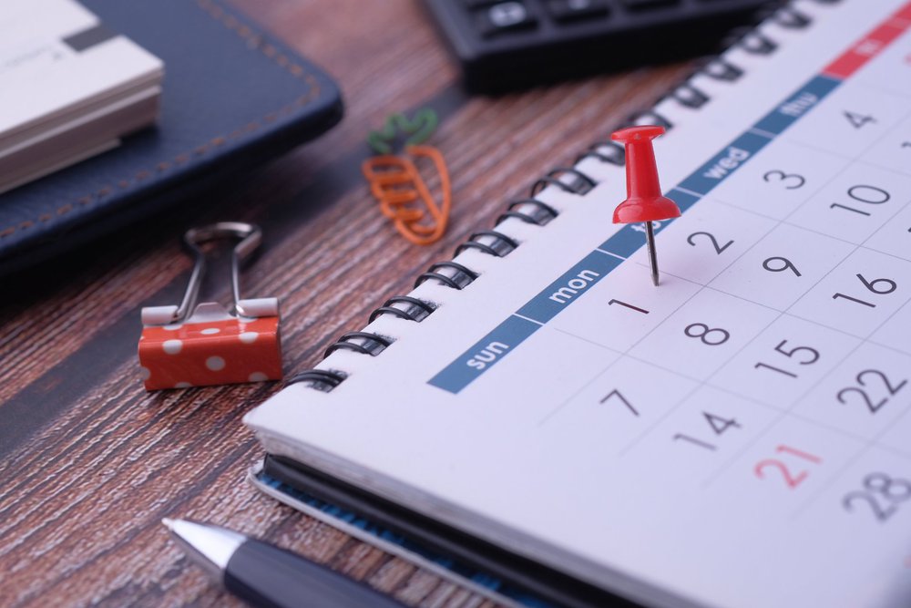 A close-up of a desk calendar with a red pushpin marking a date, alongside a binder clip, pen, and smartphone on a wooden surface.