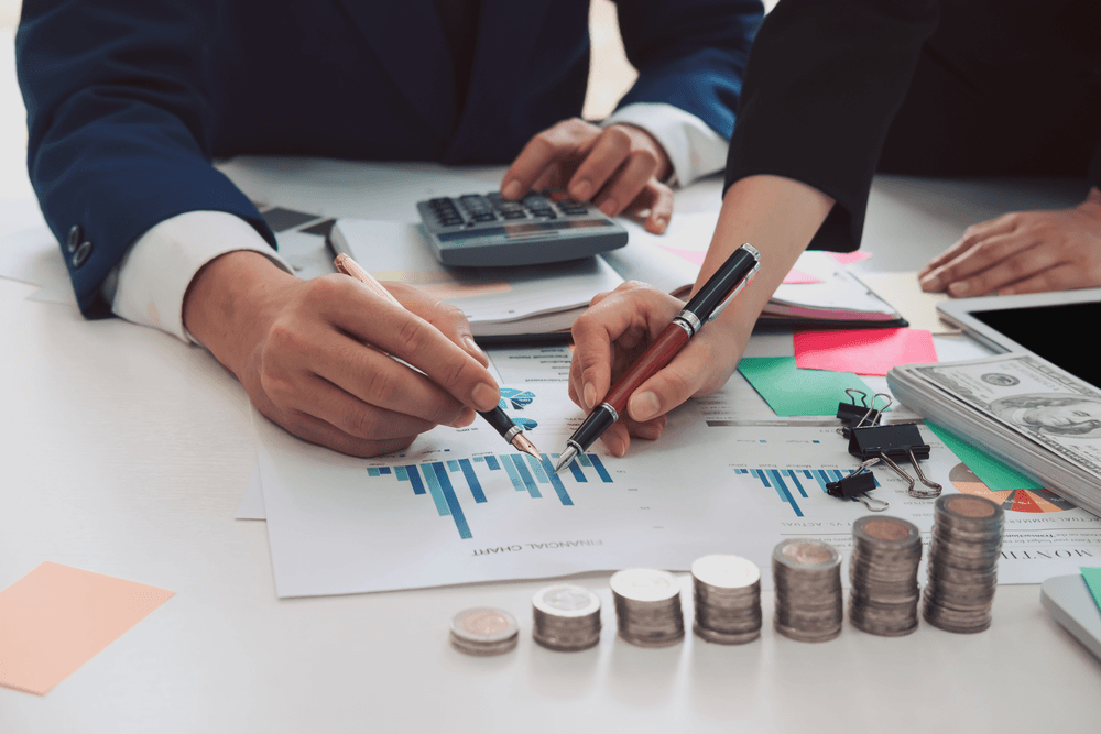 Two business professionals reviewing financial charts on a desk with stacked coins, cash, and a calculator.