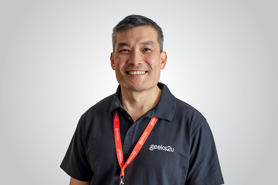 A portrait of a man smiling to camera in a navy shirt with geeks2u logo on it, wearing a red geeks2u lanyard. 