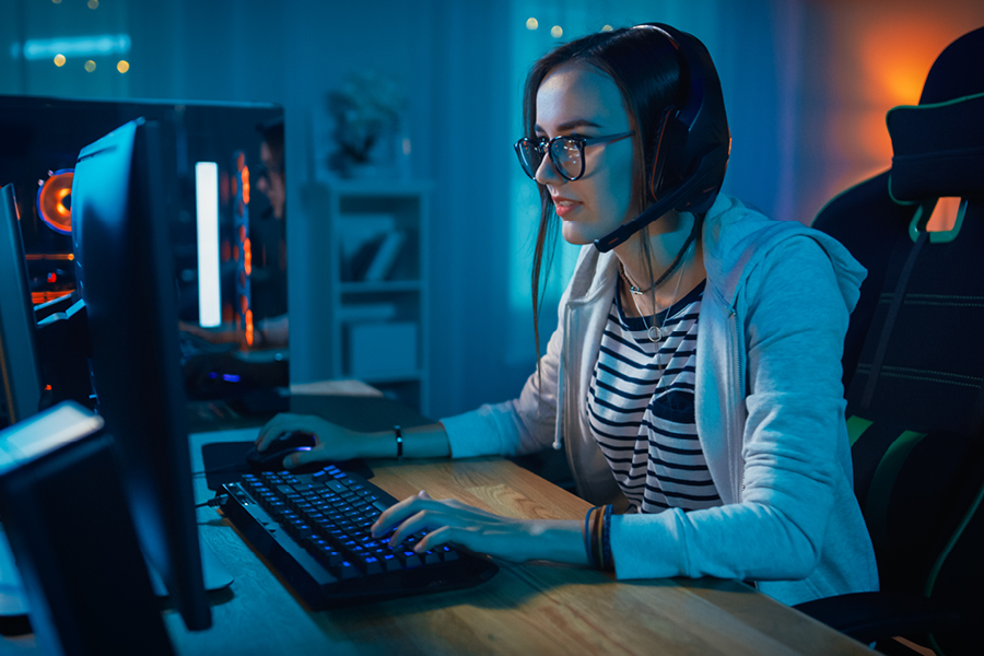 A woman sitting in front of a desktop computer, using a gaming mouse and keyboard. She’s wearing gaming headphones, black glasses, a black and white striped shirt and a grey hoodie.