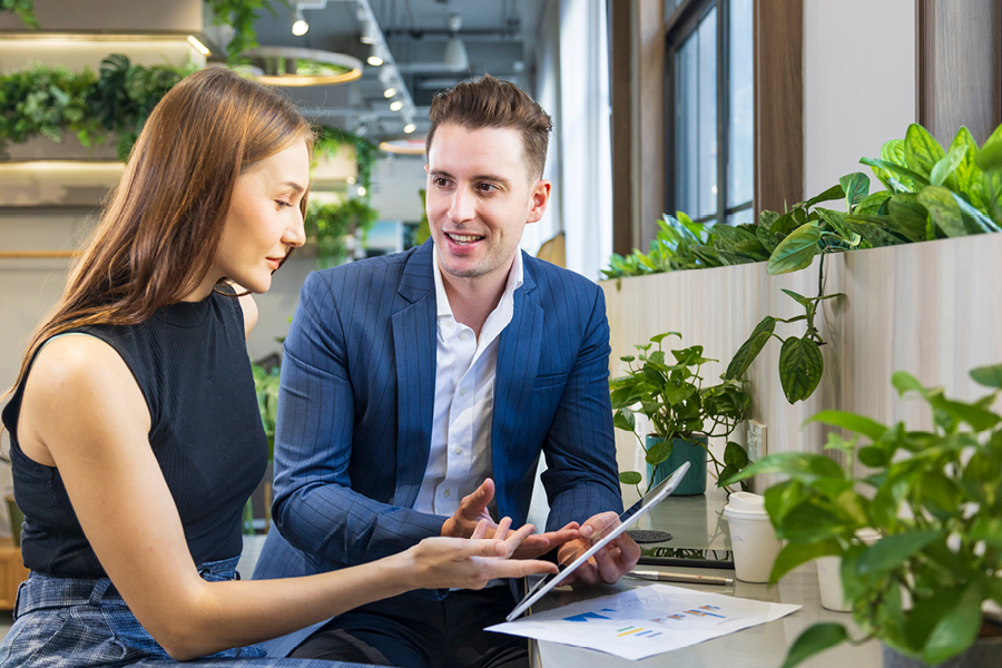 Two people collaborating on a tablet while sitting at a desk, surrounded by plants in a bright office space.