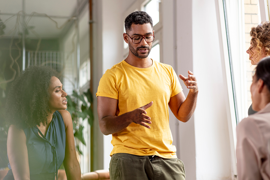 A person standing centre in an office setting is talking and moving their hands as three colleagues look towards them and listen.