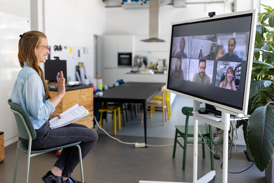 A person sitting in an office chair smiling and waving at a webcam and large screen that is showing a video call with six others who are also smiling and/or waving.