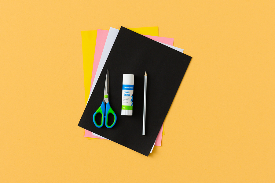 An assortment of products for making a paper pelican, arranged neatly on a yellow surface. Items include construction paper, a pencil, scissors and glue.