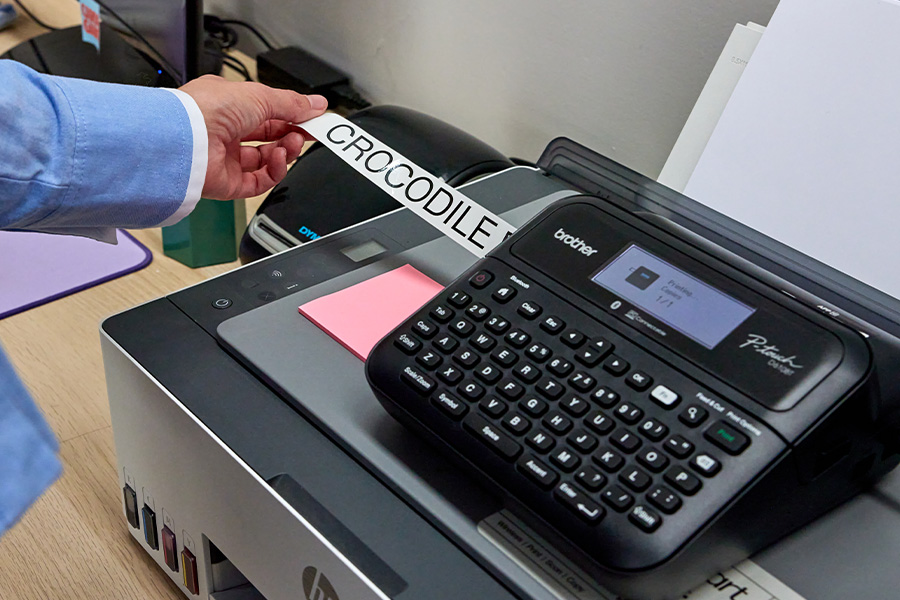 A person using a label maker to print a yarn label with the words ‘CROCODILE ROCK’. The machine sits on a desk on top of a printer and alongside sticky notes.