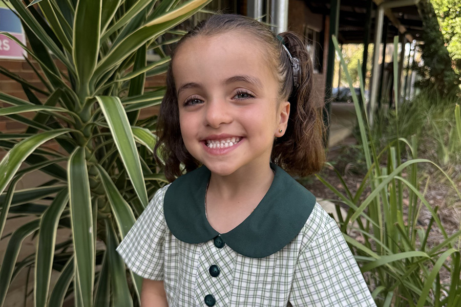 A young child with a hearing implant, smiling and standing outdoors near greenery at school.